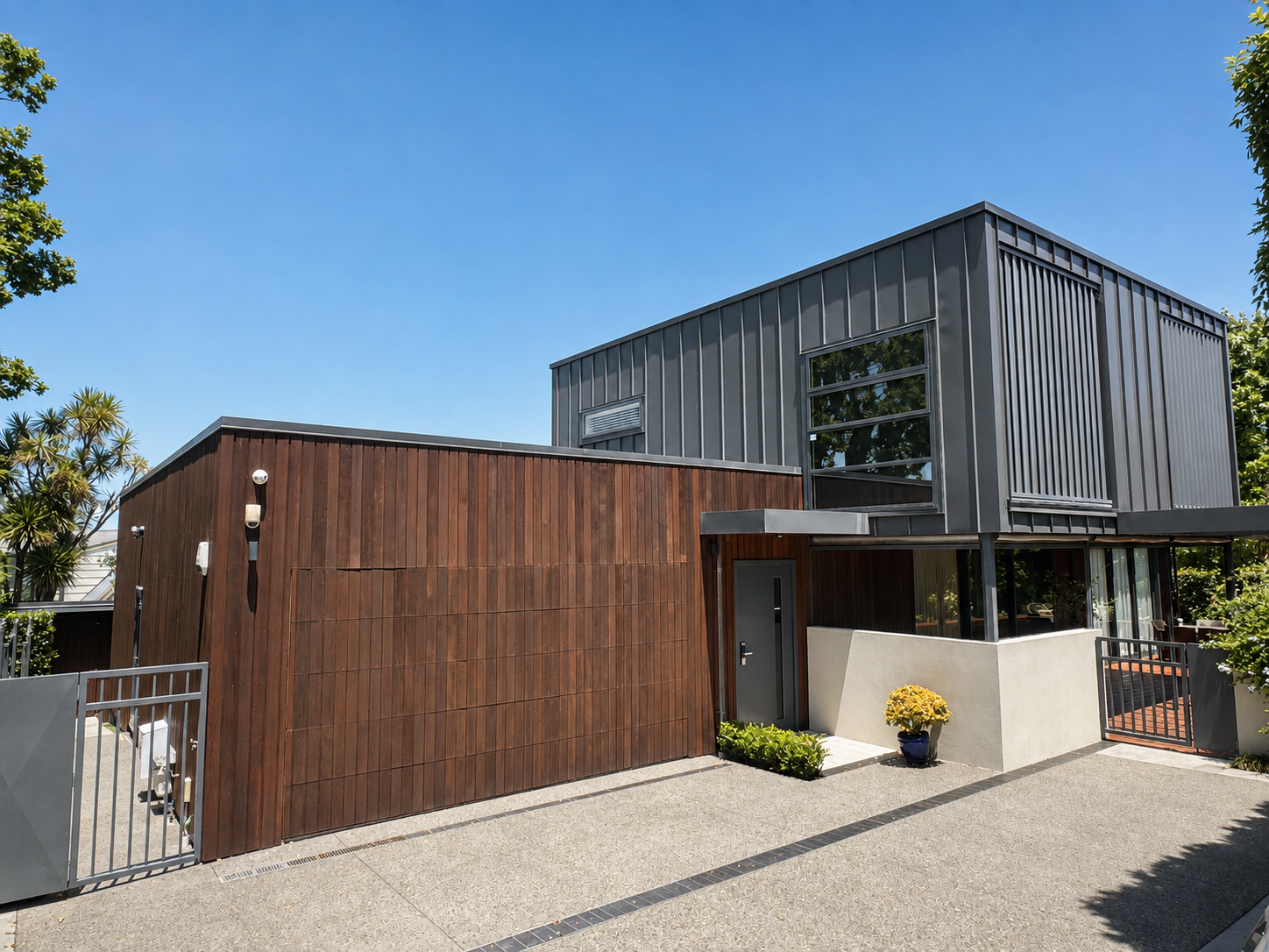 Modern house with a combination of dark wood and gray metal exterior, large windows, and a gated entrance, with a potted yellow flower outside on a sunny day.