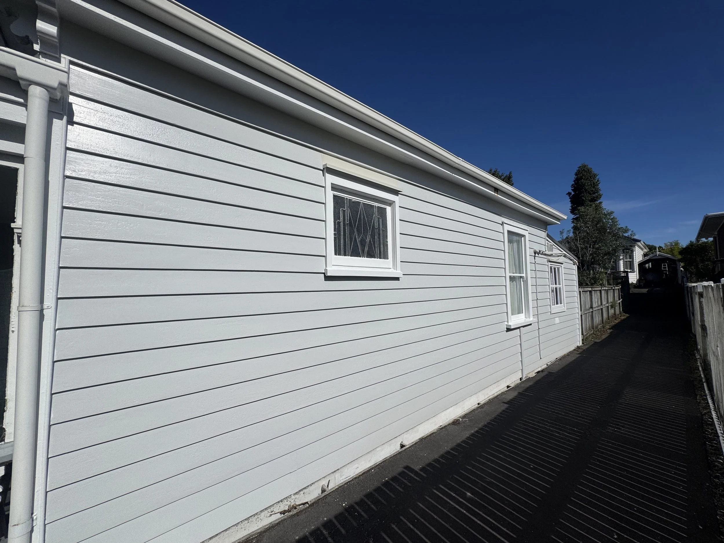 The side of a white house with horizontal siding, a window with decorative metal bars, and a downspout, under a clear blue sky with a tree in the background.