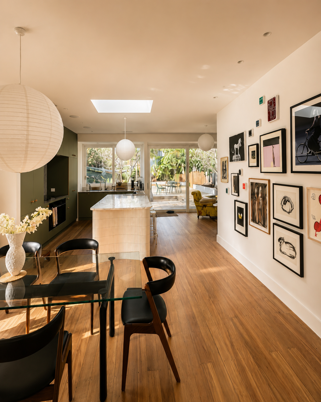 Interior of a modern dining area and kitchen with wooden flooring, a glass dining table, black chairs, a white wall with framed artwork, pendant lights, and a view of an outdoor patio with garden furniture.