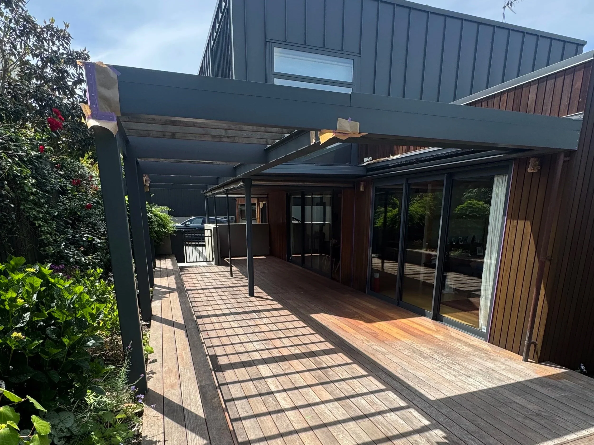 Modern house with large glass windows, wooden deck, and attached pergola under construction with building materials visible, surrounded by green trees and plants.