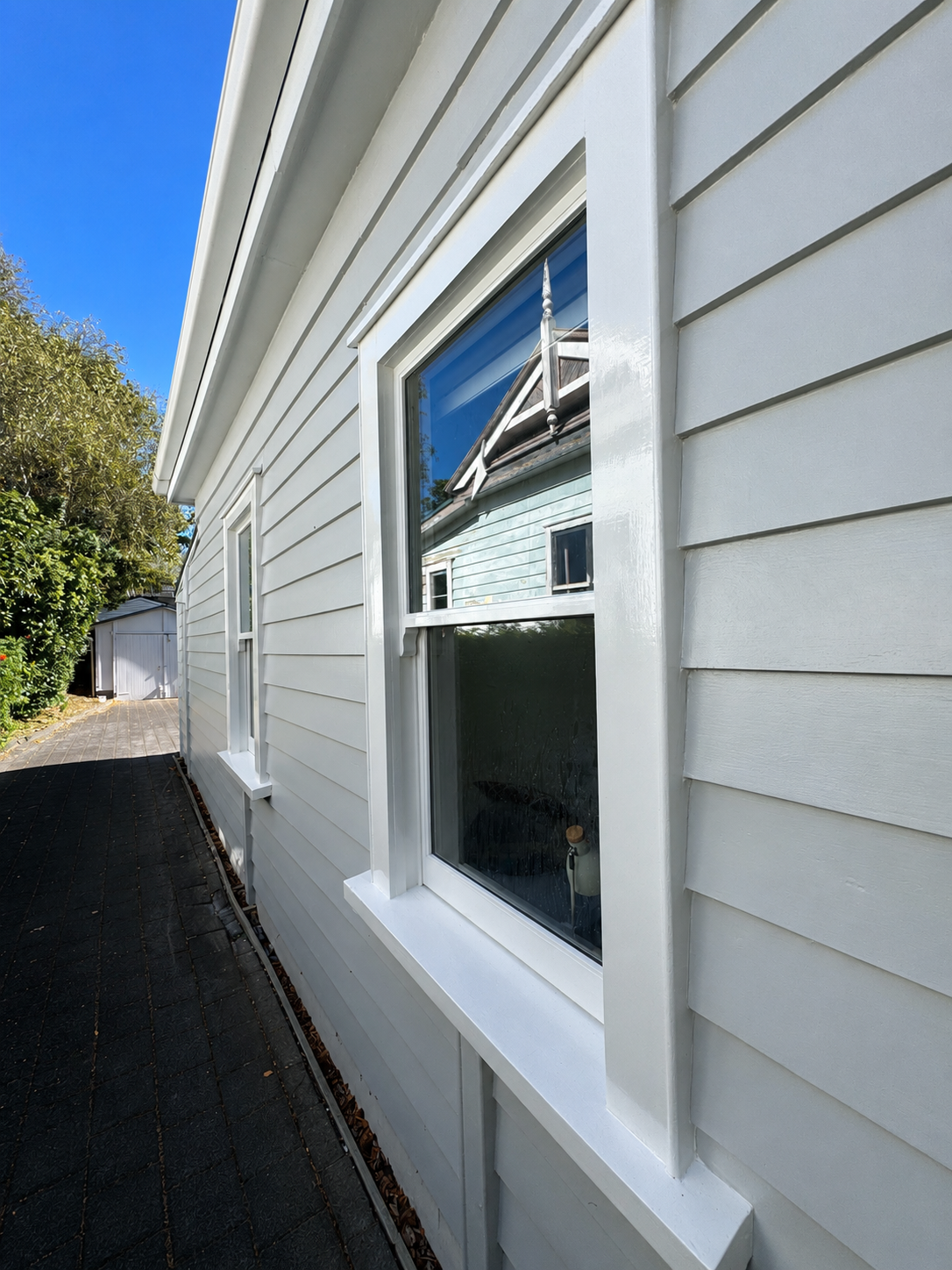 Side view of a white house with vinyl siding, showing two double-pane windows with white trim, a narrow walkway and a garden shed in the distance on a sunny day.