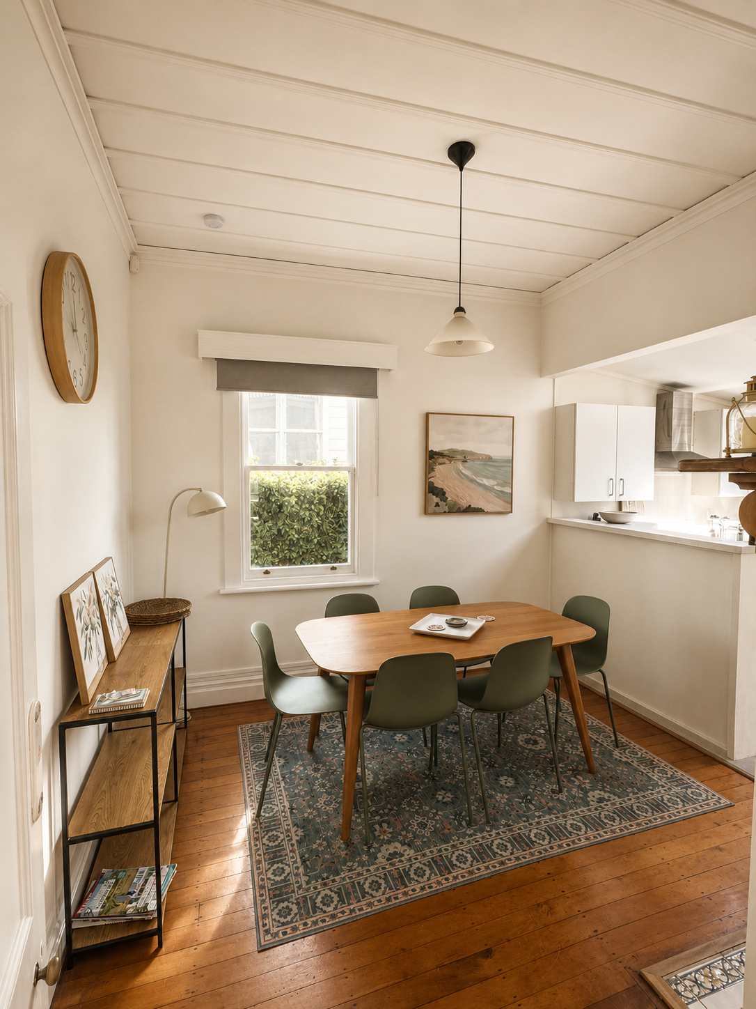 Dining room with a wooden table and six green chairs, a window with plants outside, framed artwork, a bookshelf with magazines, and a patterned rug on wooden flooring.