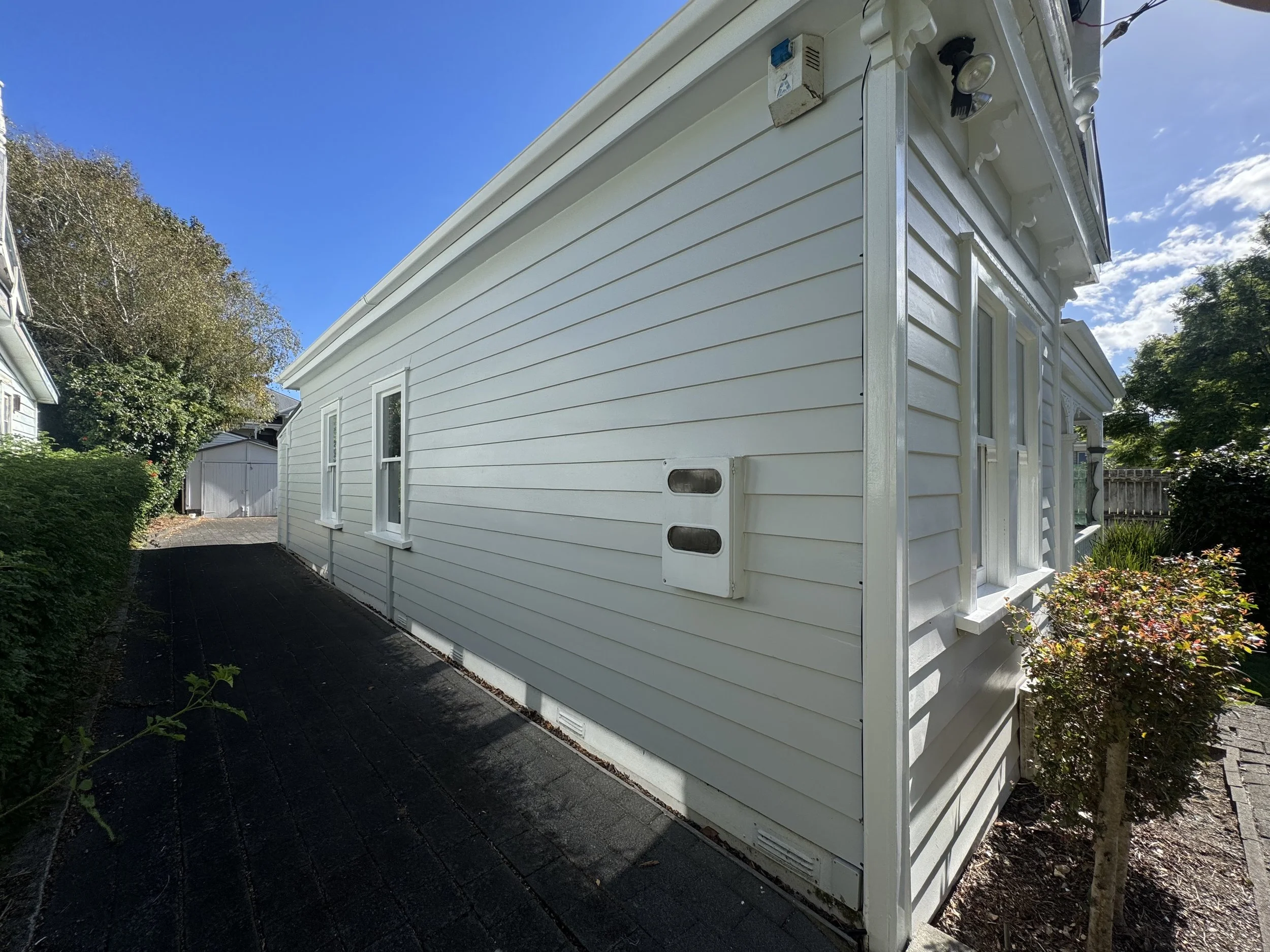 Side view of a white house with vinyl siding, small windows, and outdoor lighting. There are trees, a hedge, and a driveway with a shed in the background under a blue sky with some clouds.