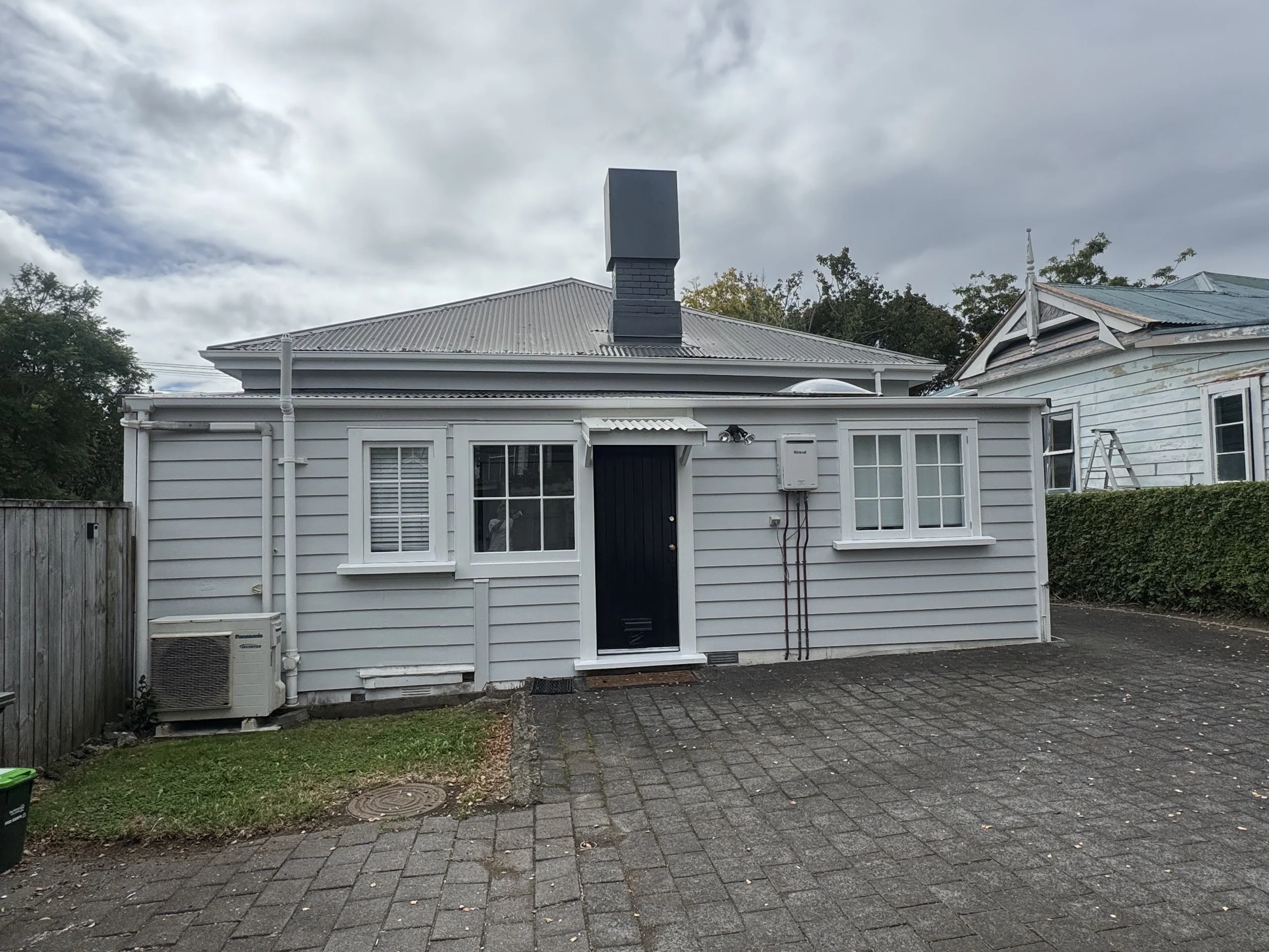 The back of a white house with a metal roof and a black chimney in a cloudy sky. There is a small window and a door on the house's rear wall, with an air conditioning unit and various piping attached. A small patch of grass, a paved driveway, and a w