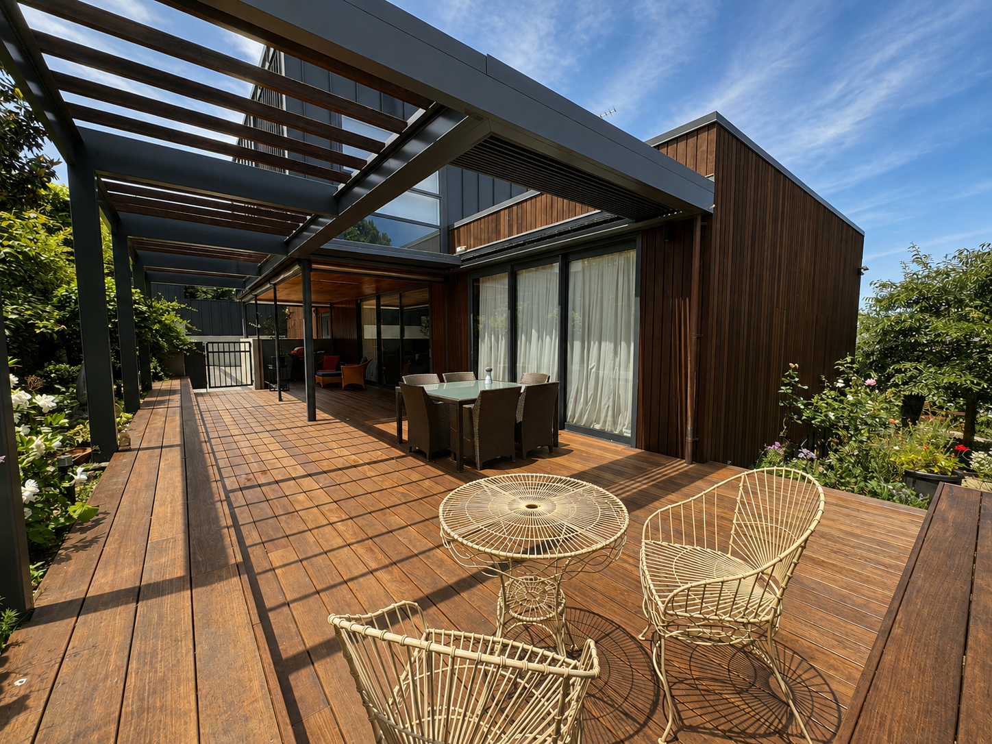 Modern house with a large wooden deck, outdoor furniture including a round table with chairs and a lounge area, partially covered by a pergola with wooden beams, surrounded by greenery and flowers, under a partly cloudy sky.