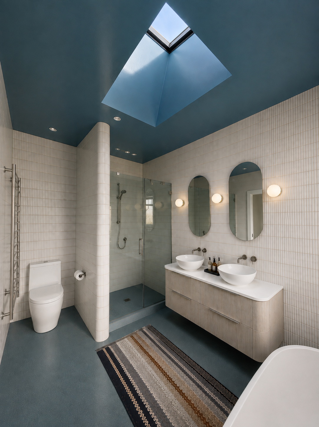 Modern bathroom with blue ceiling, skylight, white tiled walls, glass shower enclosure, toilet, double sinks with oval mirrors, wall-mounted lights, striped rug, and a freestanding bathtub.