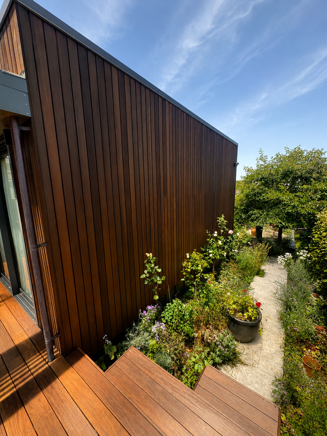 Wooden house exterior with a garden path, potted plants, and flowers facing a clear blue sky.