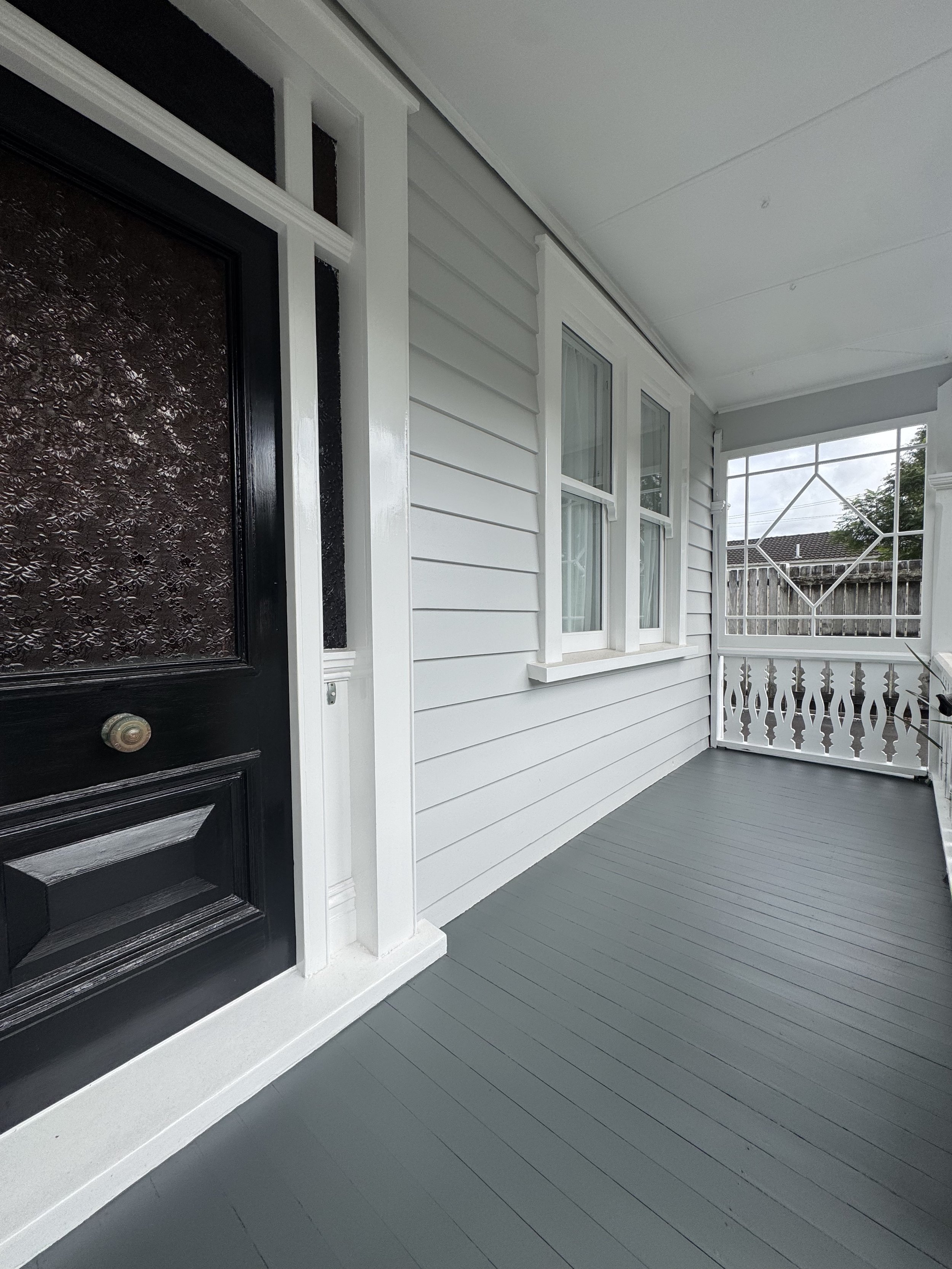 Front porch of a house with white siding, double-pane windows, a dark door with textured glass, and a decorative white railing on the porch.