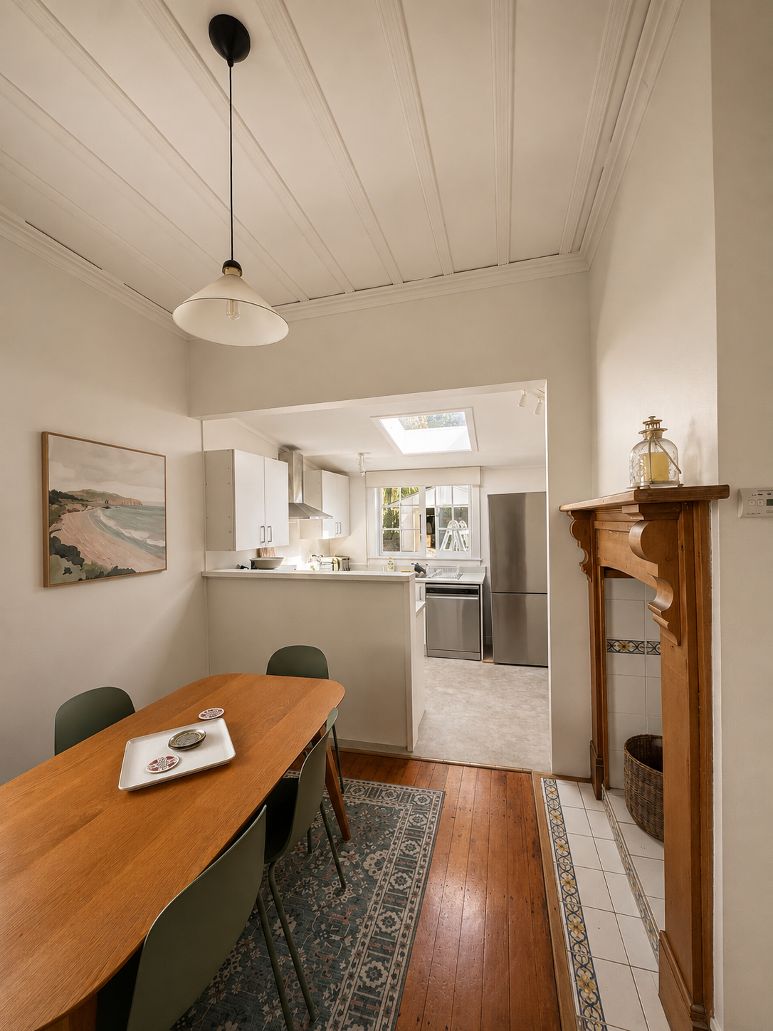A dining area with a wooden table and four green chairs, a decorative carpet, and a white ceiling with a hanging lamp. The kitchen is visible in the background with white cabinets, a stainless steel refrigerator, and a window letting in natural light