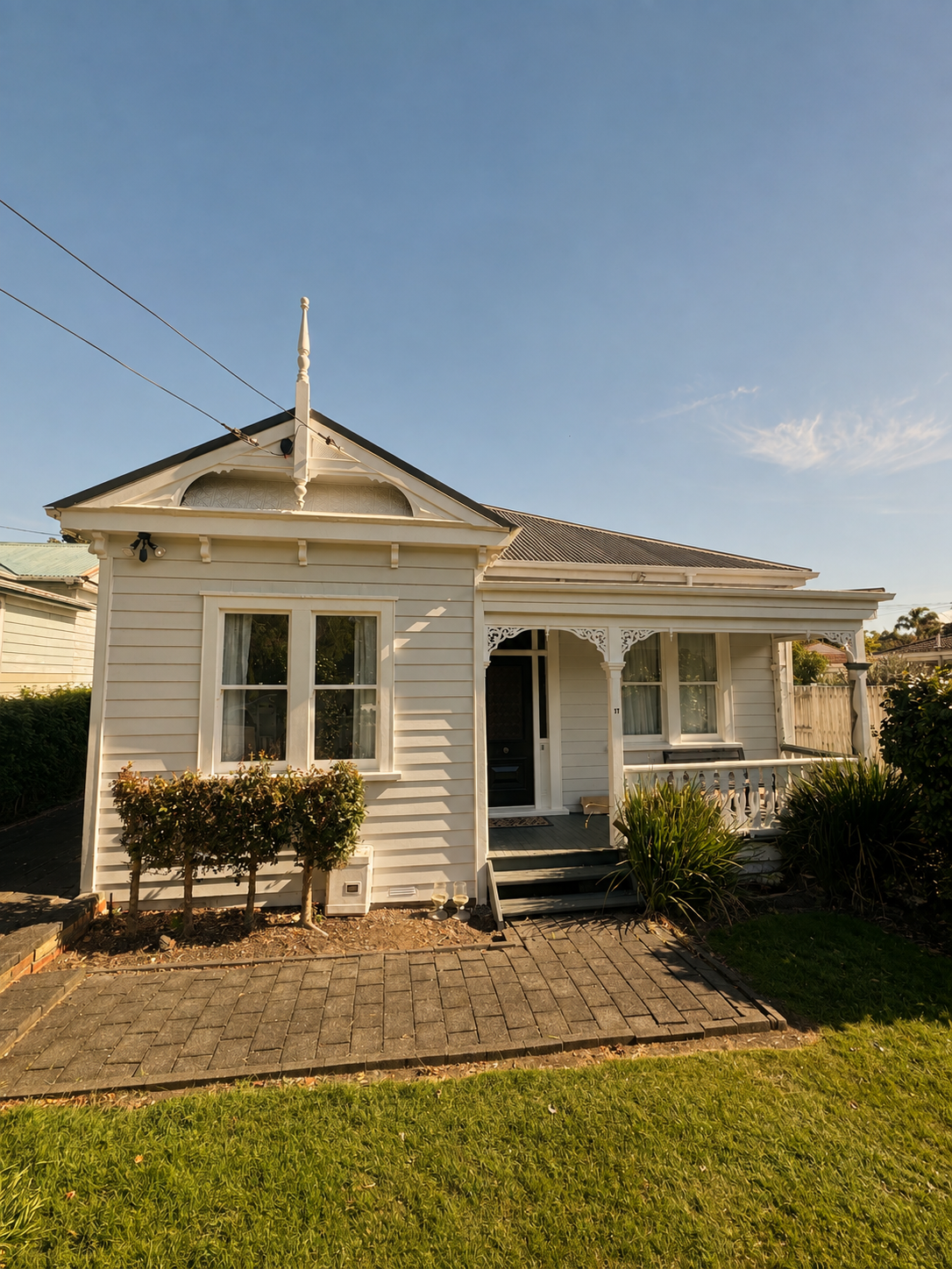 Front view of a small, white wooden house with decorative trim and a front porch, surrounded by a lawn and shrubbery under a clear blue sky.
