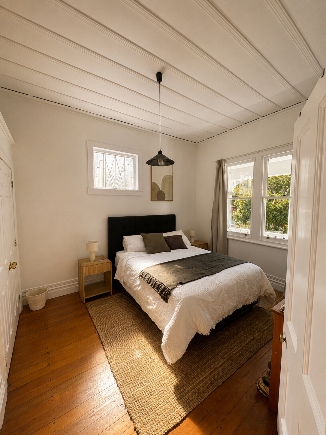 A cozy bedroom with a wooden floor, white walls, a bed with a black headboard, and a beige and white bedspread. There are two nightstands with lamps, a small window with decorative bars, a larger window with curtains, and a small piece of abstract ar