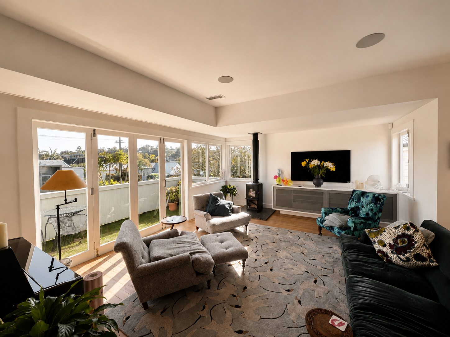 Living room with large windows, various chairs, a sofa, a TV, potted plants, and a patterned rug.