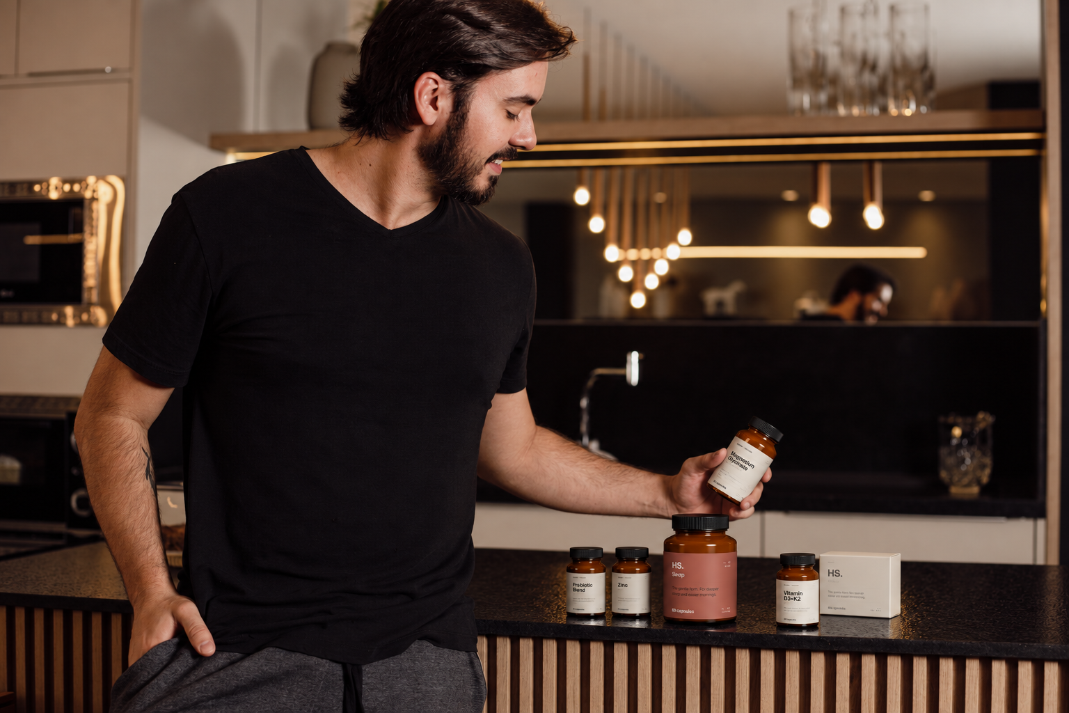 A man in a black t-shirt and gray pants standing in a modern kitchen, holding a bottle of supplement while looking at it. There are several supplement bottles and boxes on the kitchen counter.