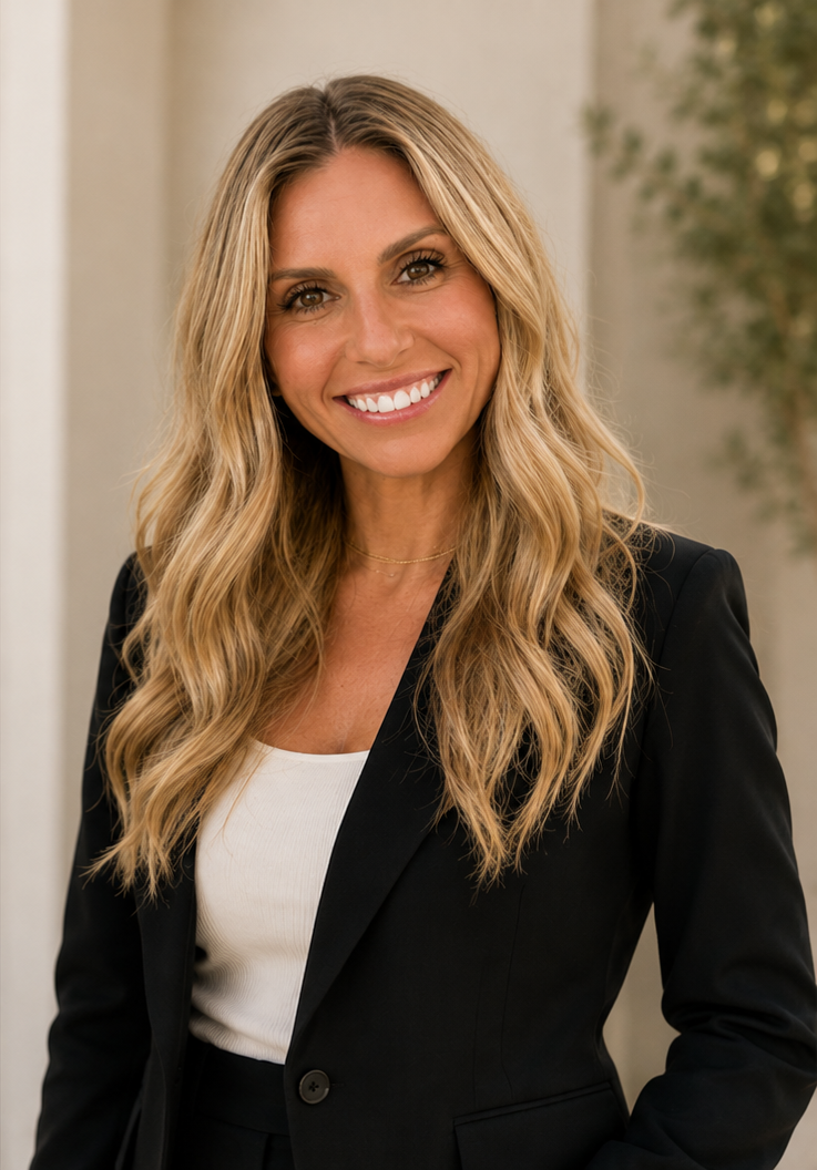 A woman with long blonde hair smiling at the camera, dressed in a black blazer and white top, standing indoors with a blurred background.