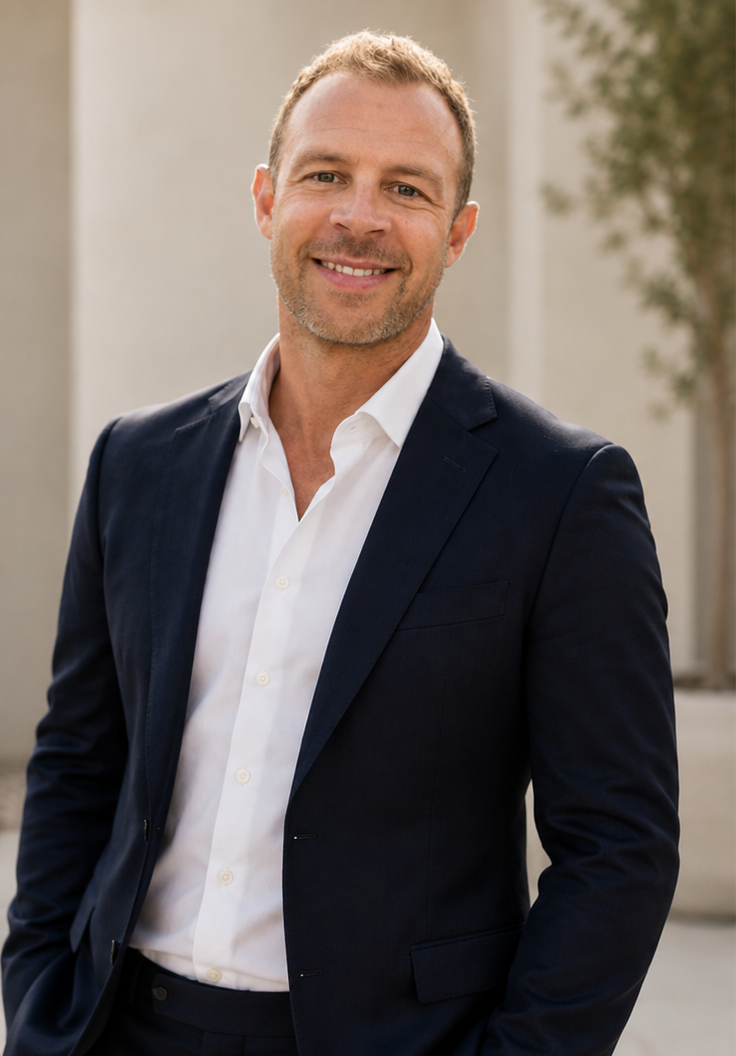 A smiling man in a white shirt and dark blazer standing outdoors with a blurred background of trees and a building.