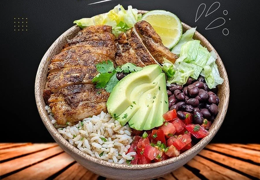 A bowl of grilled chicken, rice, avocado slices, chopped tomatoes, black beans, lettuce, and lemon wedge.