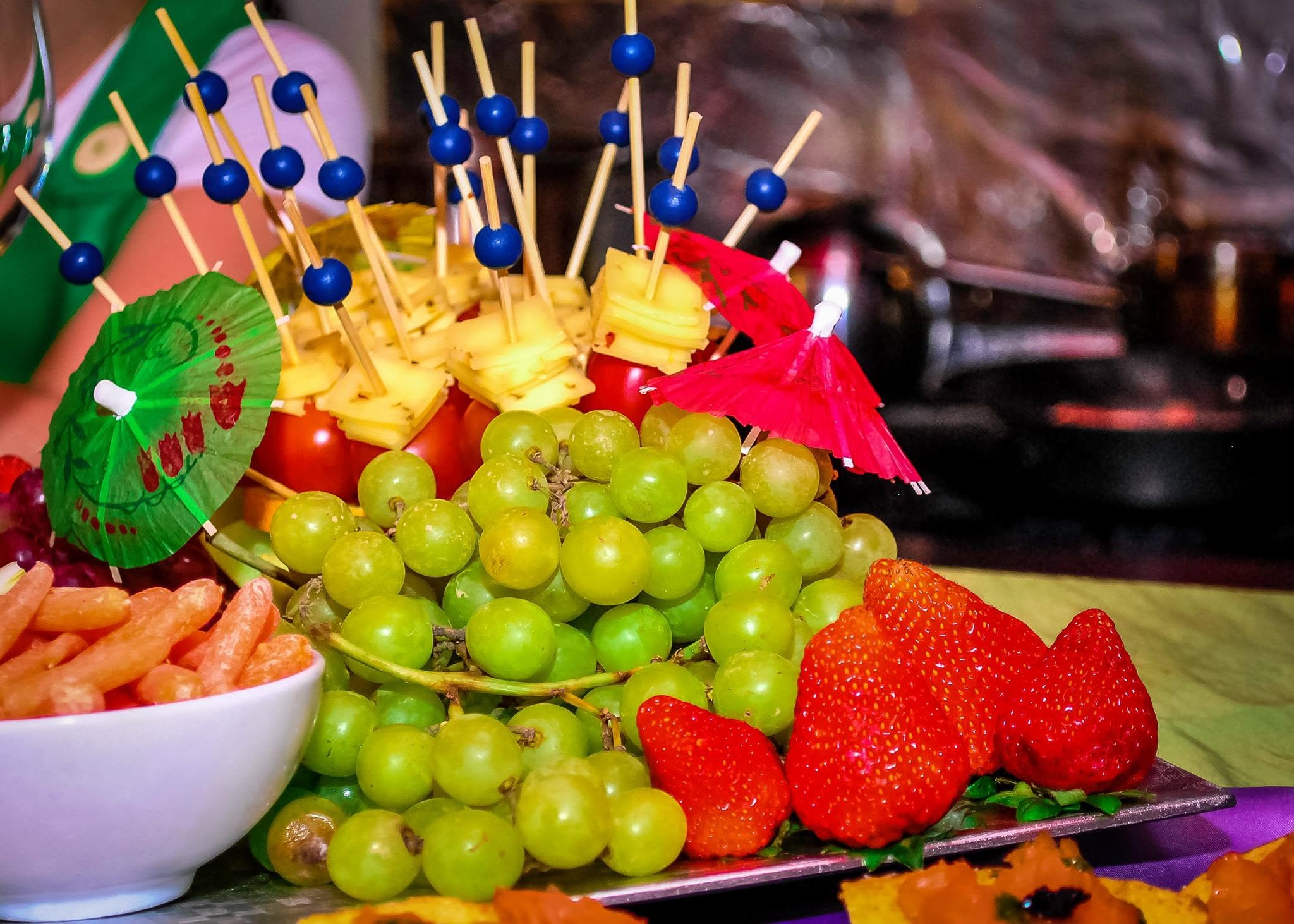 Close-up of a colorful fruit display, including green grapes, strawberries, pineapple slices on skewers with small umbrellas, and other assorted fruits.