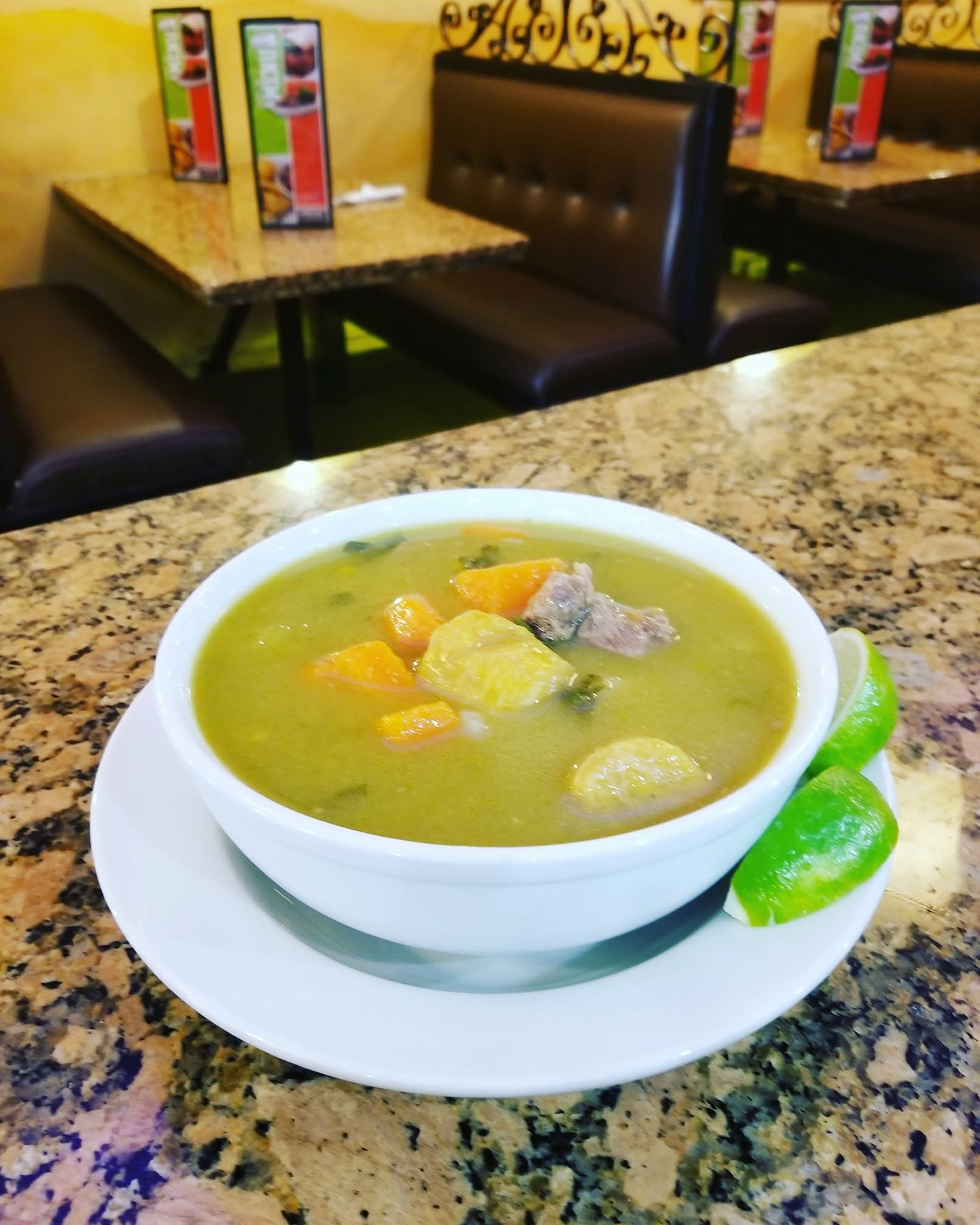 A bowl of green vegetable soup with chunks of carrots and meat, garnished with lime wedges on a white plate, on a granite countertop in a restaurant.