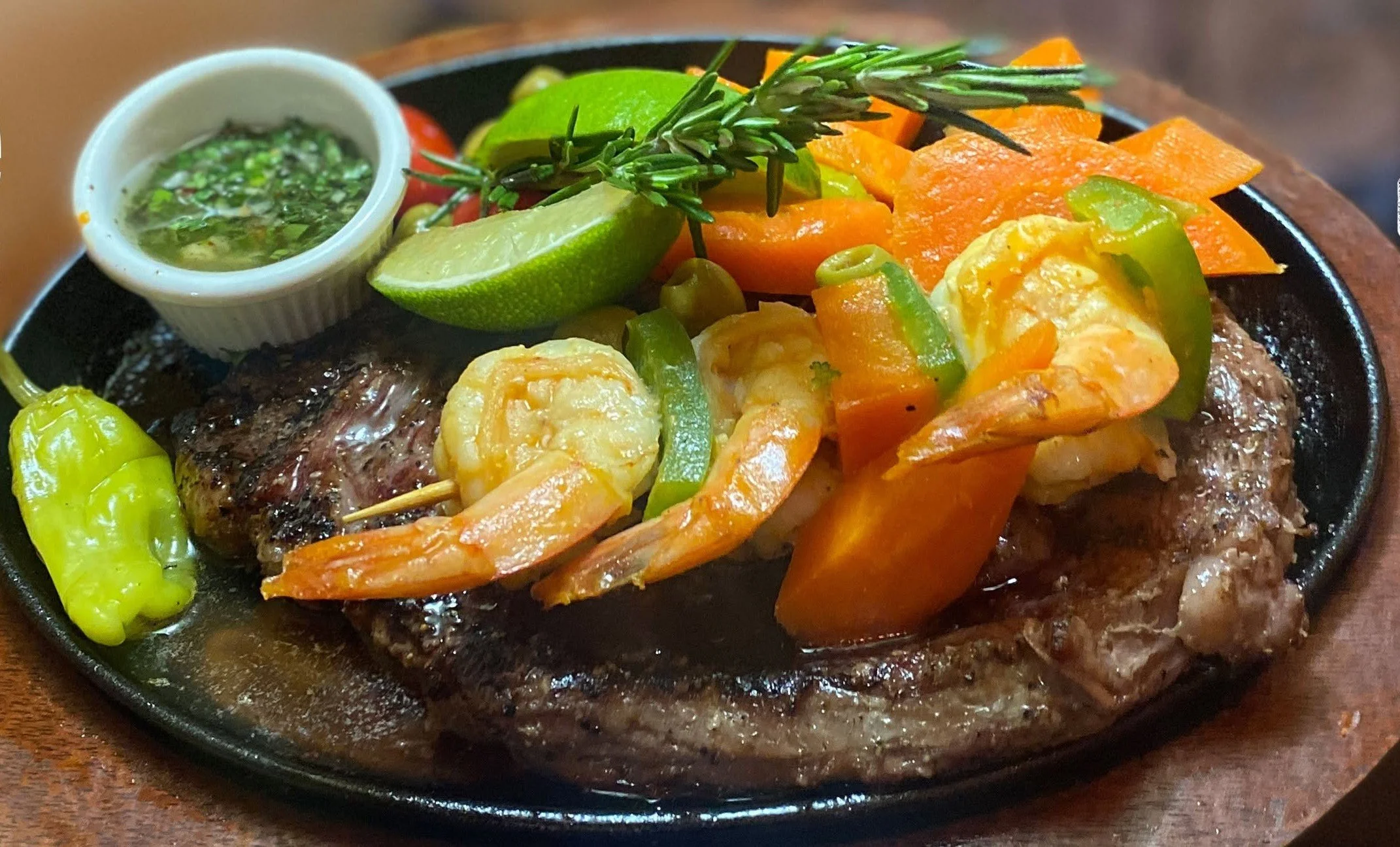 A sizzling plate of food featuring grilled steak topped with grilled shrimp, garnished with a sprig of rosemary, lime, and green chili peppers, accompanied by a small bowl of green sauce and chopped vegetables.
