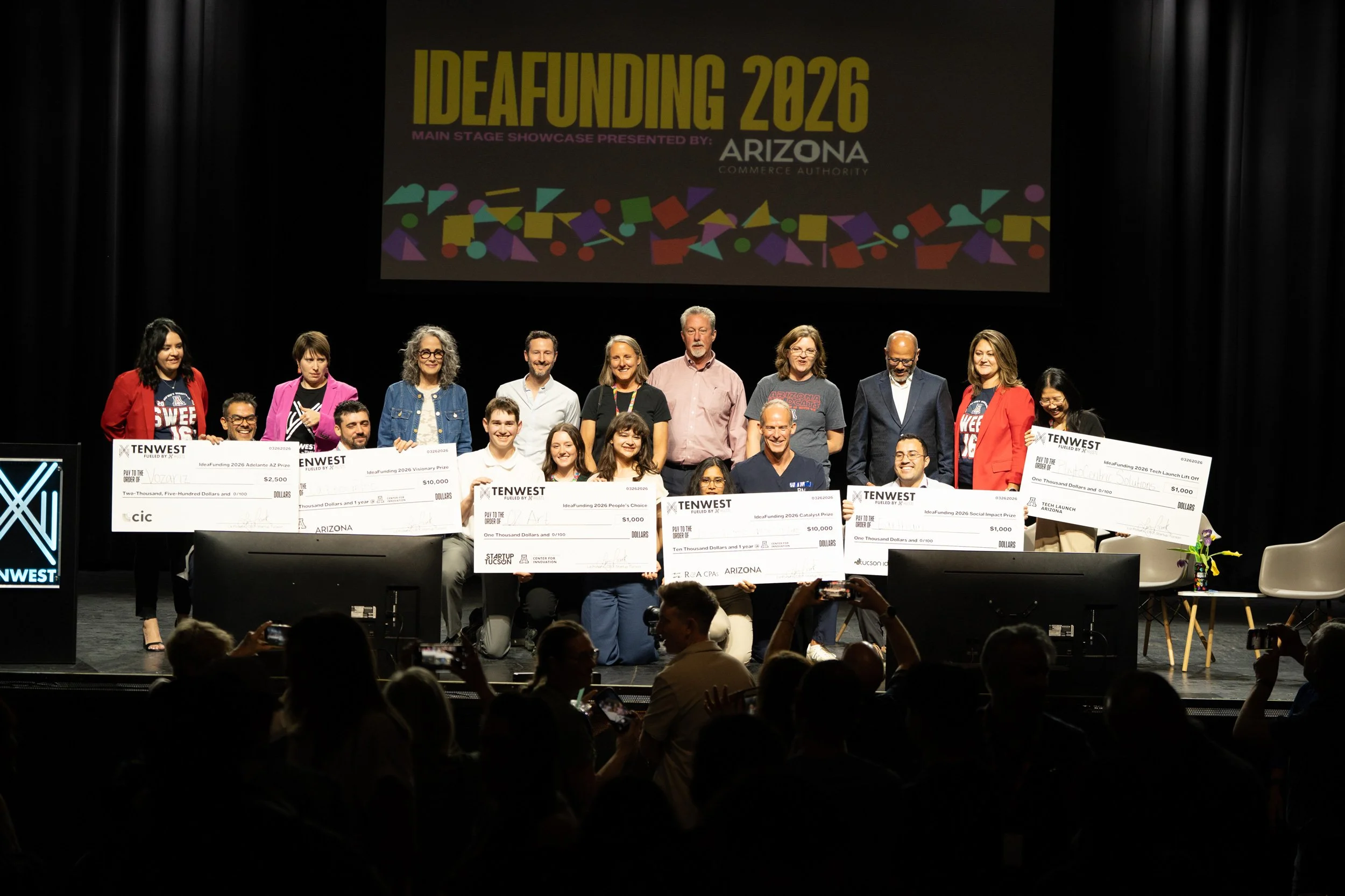 Group of people on stage holding large checks at an event called IDEAFUNDING 2026 in Arizona, with a large screen displaying event details behind them.