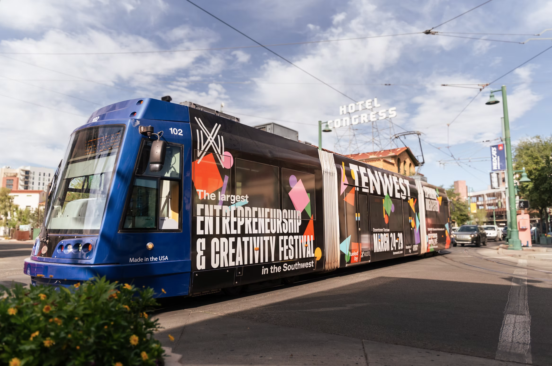 A colorful tram decorated with advertisements for an entrepreneurship and creativity festival, with the words 'The largest ENTREPRENEURSHIP & CREATIVITY FESTIVAL' visible. In the background, there is a sign that reads 'HOTEL CONGRESS' against a partly cloudy sky, and some buildings and trees are also visible.