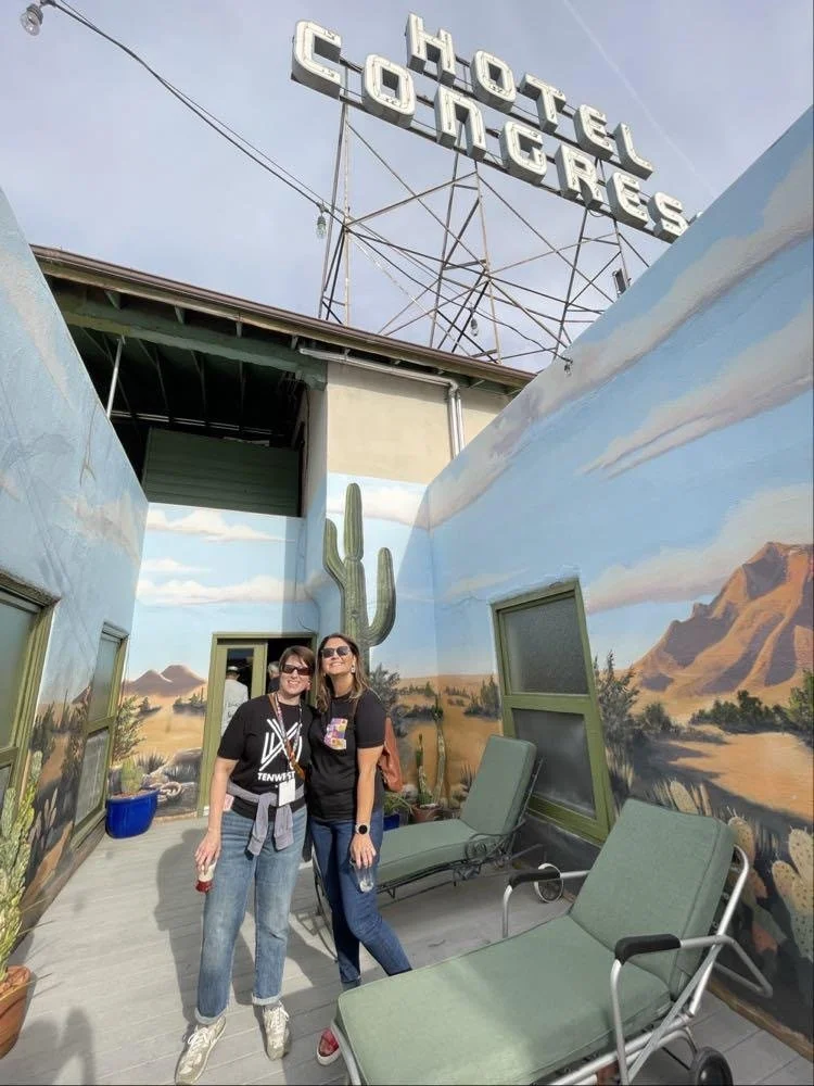 Two women standing on a rooftop with desert mountain mural walls and cactus paintings, with lounge chairs and a large sign that reads 'Hotel Congress' in the background.