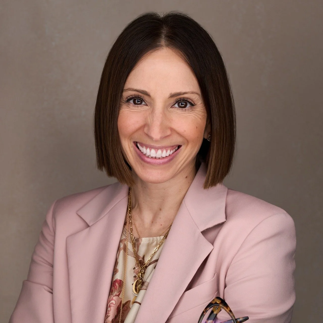 A woman with shoulder-length brown hair smiling, wearing a light pink blazer and patterned blouse, standing against a plain background.