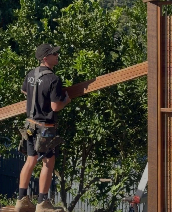 A person wearing a black cap, sunglasses, and a black work shirt with 'SQUAD' on the back, is holding a wooden plank and working on a construction project outside near a tree and a partially built wooden structure.