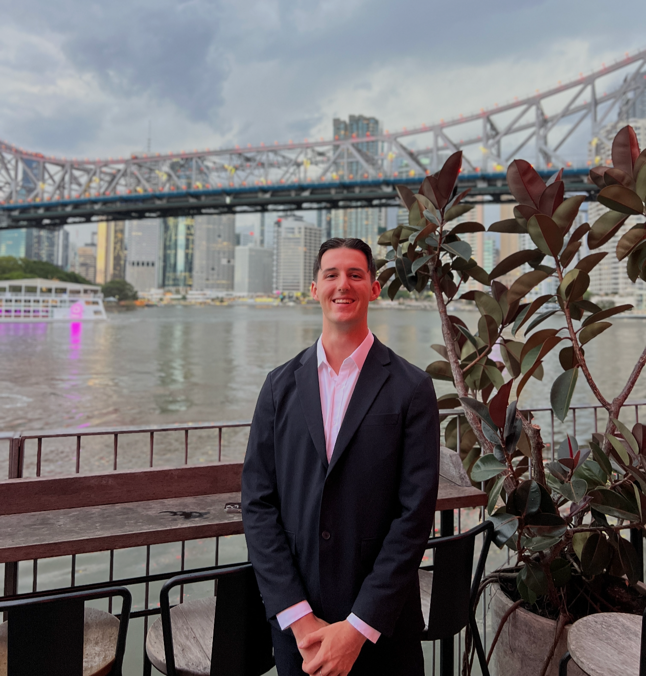 A man in a black suit, white shirt, smiling, standing outdoors near a river, with a city skyline and a bridge in the background during the evening or late afternoon.