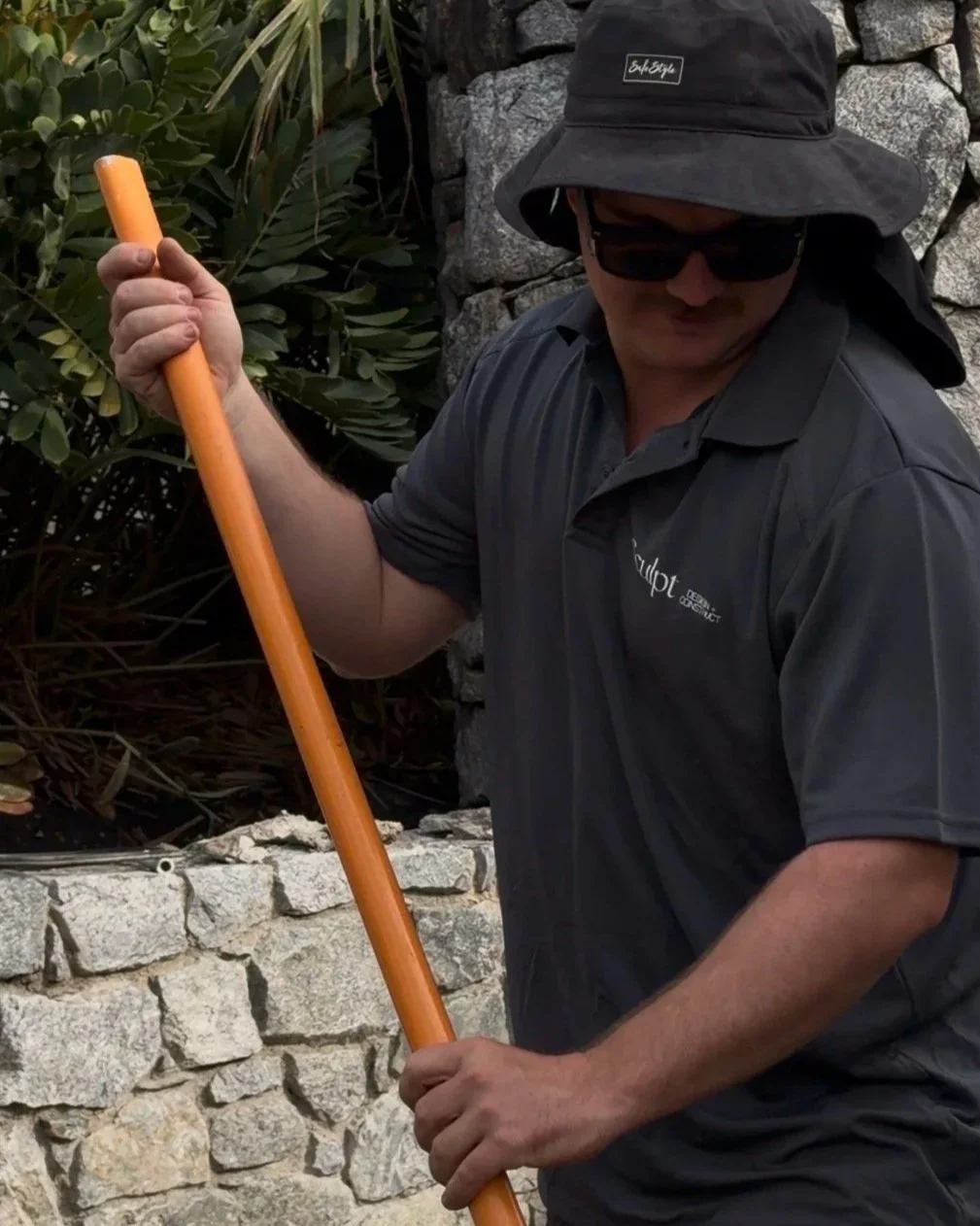Man wearing a black sun hat, sunglasses, and a dark gray shirt holding a long orange gardening tool or stick, standing outdoors near a stone wall with green foliage.
