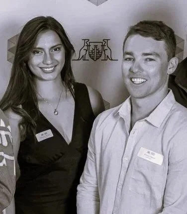 A young woman and a young man smiling and posing for a photo at an event, with a backdrop that has abstract geometric shapes and a tree sculpture in the background.