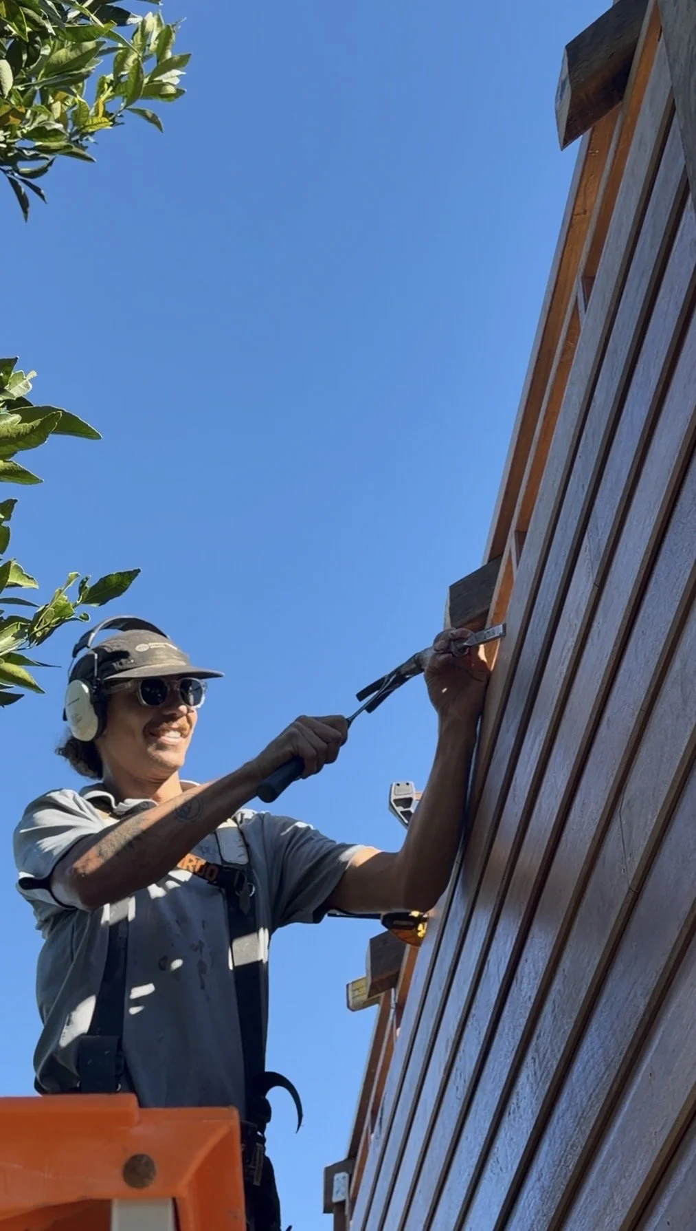 A man with sunglasses, a hat, and headphones trimming or working on a wooden exterior wall of a building with a tool, standing on an orange lift against a clear blue sky with some green leaves visible.