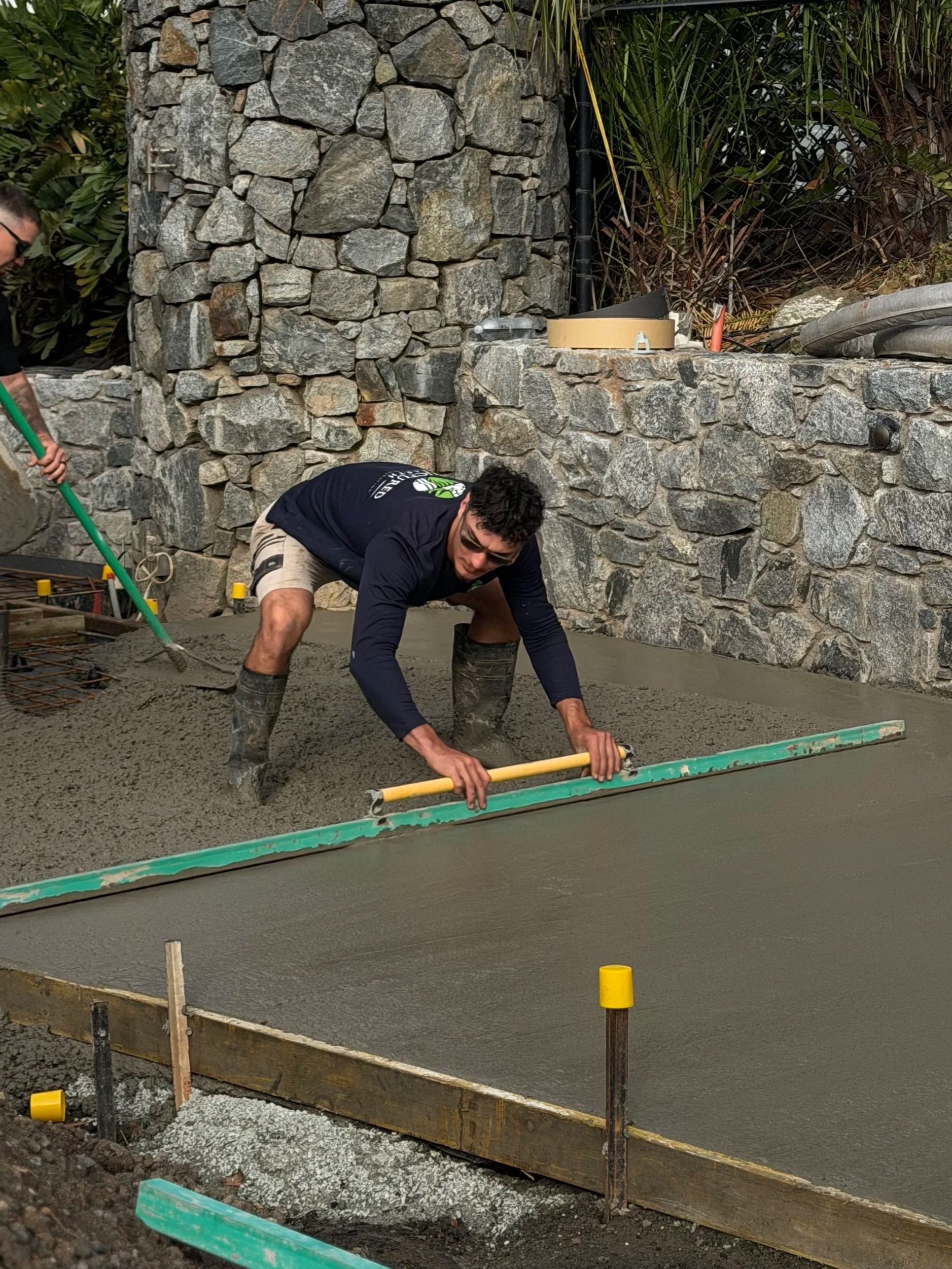 Construction workers pouring and smoothing concrete on a construction site, with a stone wall and construction tools visible in the background.