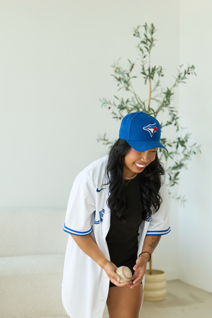 Nicole Coronel in a Toronto Blue Jays cap and baseball jersey holding a baseball indoors, supporting her favorite baseball team.
