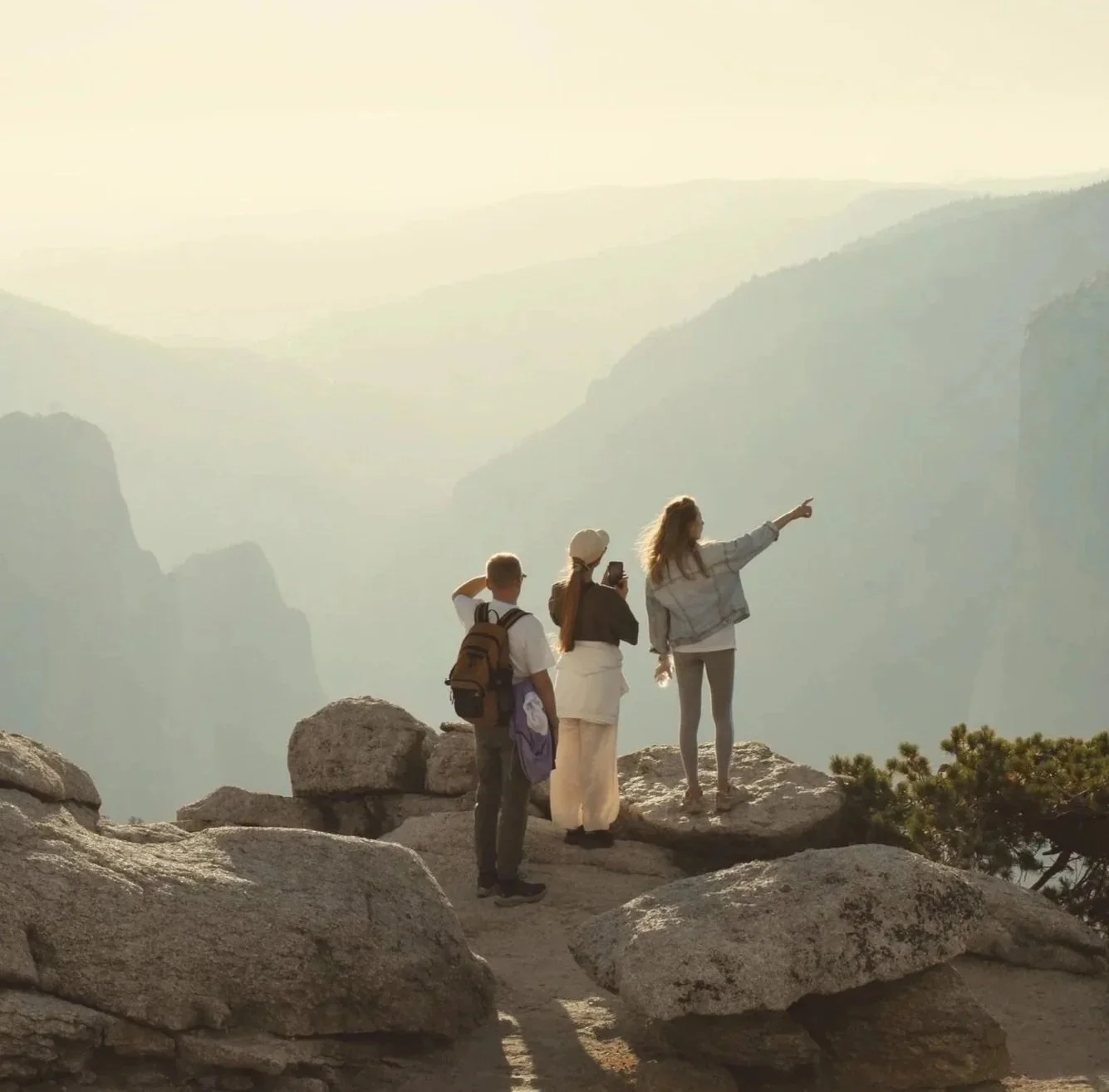 Three tourists standing on rocks at a mountain overlook, with a woman pointing at the scenic view while two others observe, against a hazy mountain backdrop.