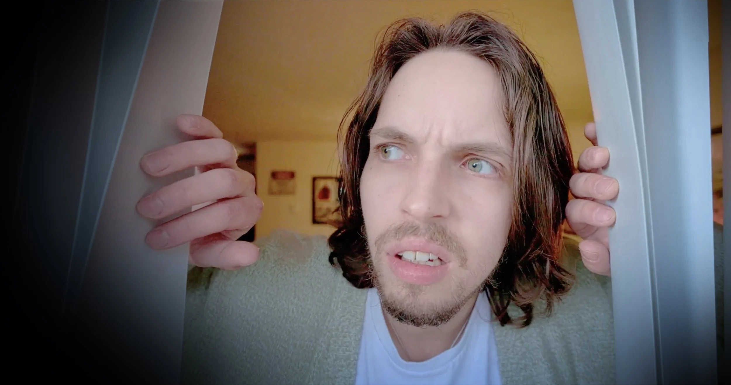 A man with long brown hair and a beard looks through a doorway, holding the door frame with both hands, with a slightly confused or surprised expression.