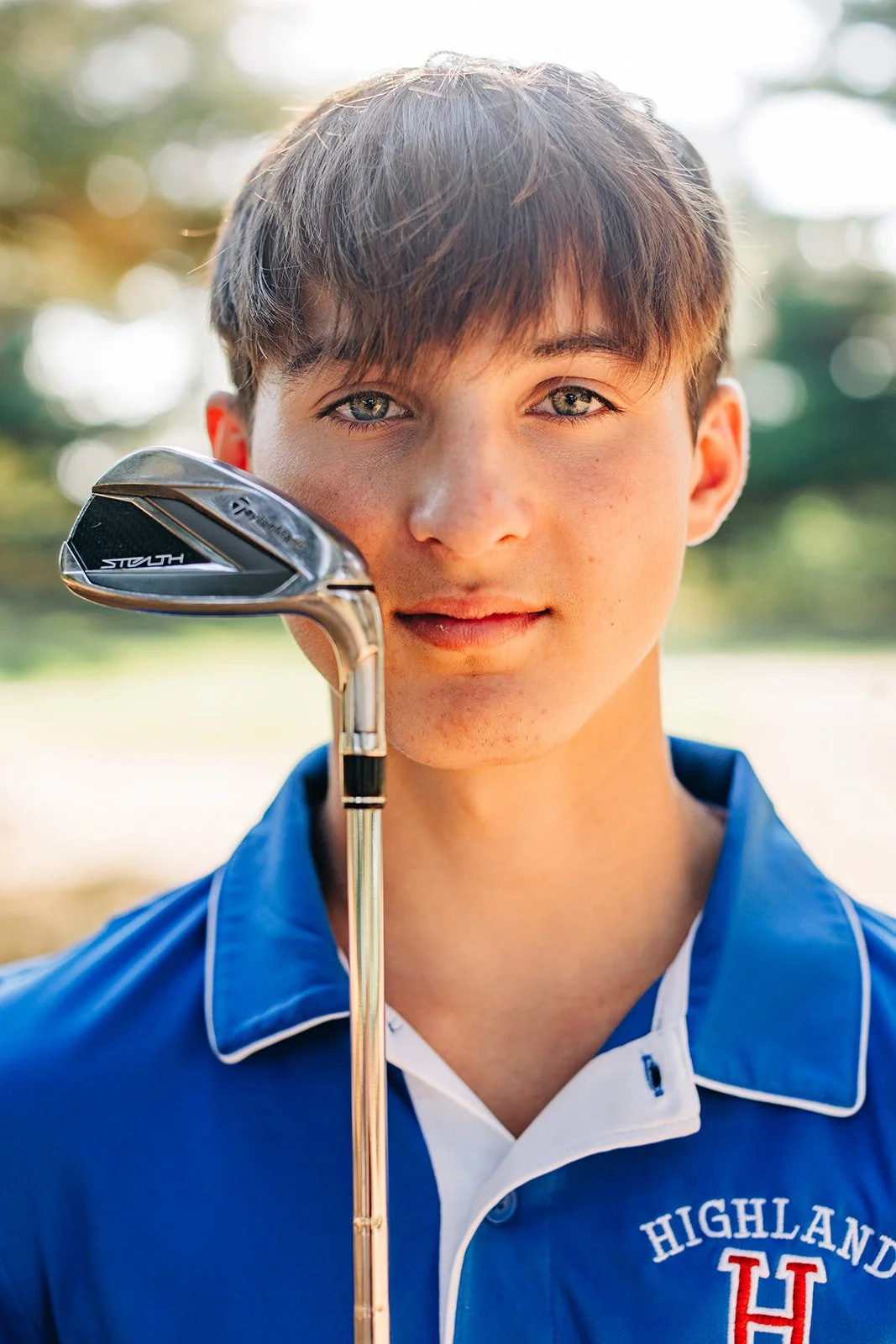 A young man with light brown hair holds a golf club, standing outdoors with blurred trees and greenery in the background. He is wearing a blue polo shirt with white accents and the word 'Highland' and the letter 'H' embroidered on it.