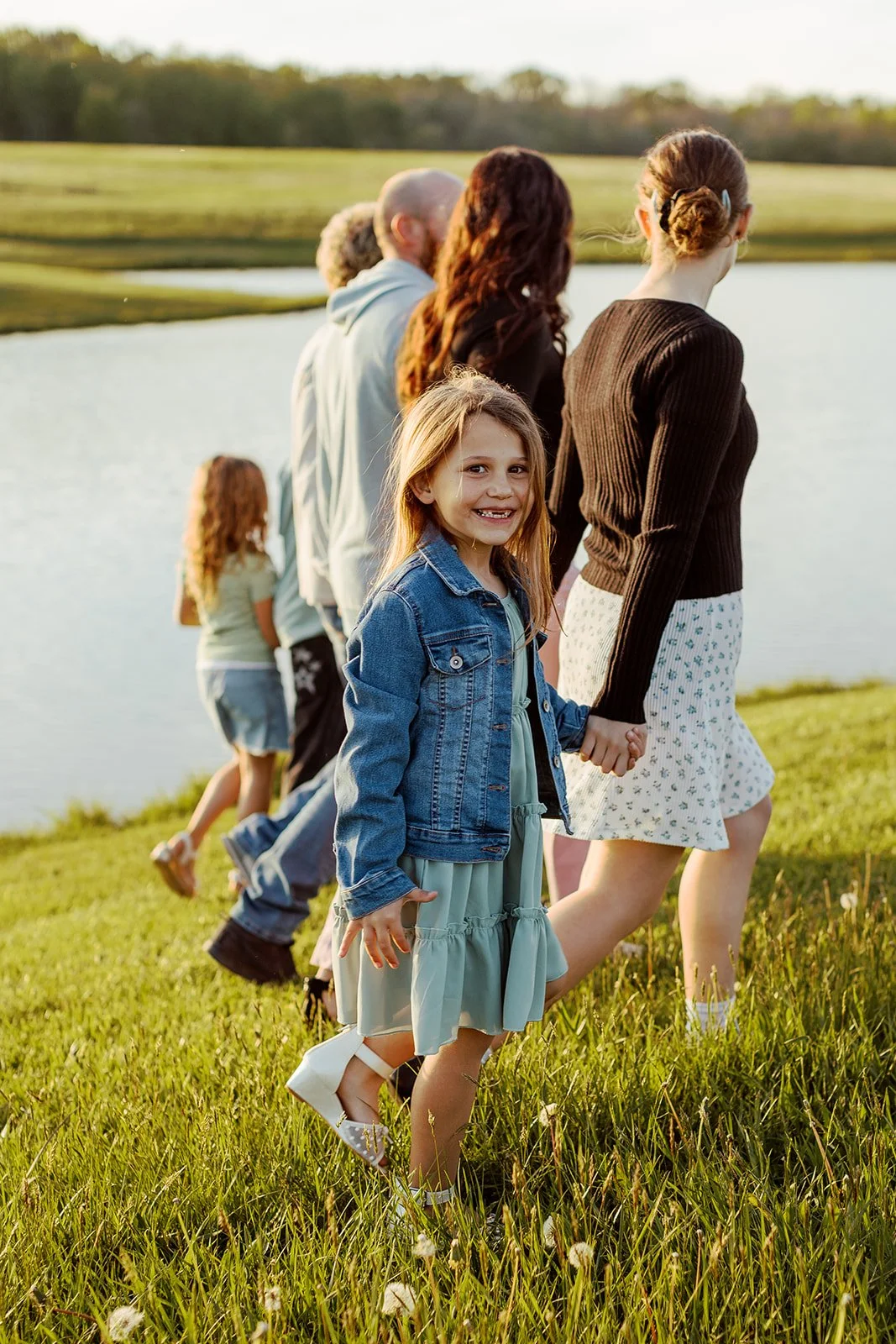 A group of people, including children and adults, walking hand in hand along a grassy area near a body of water at sunset.