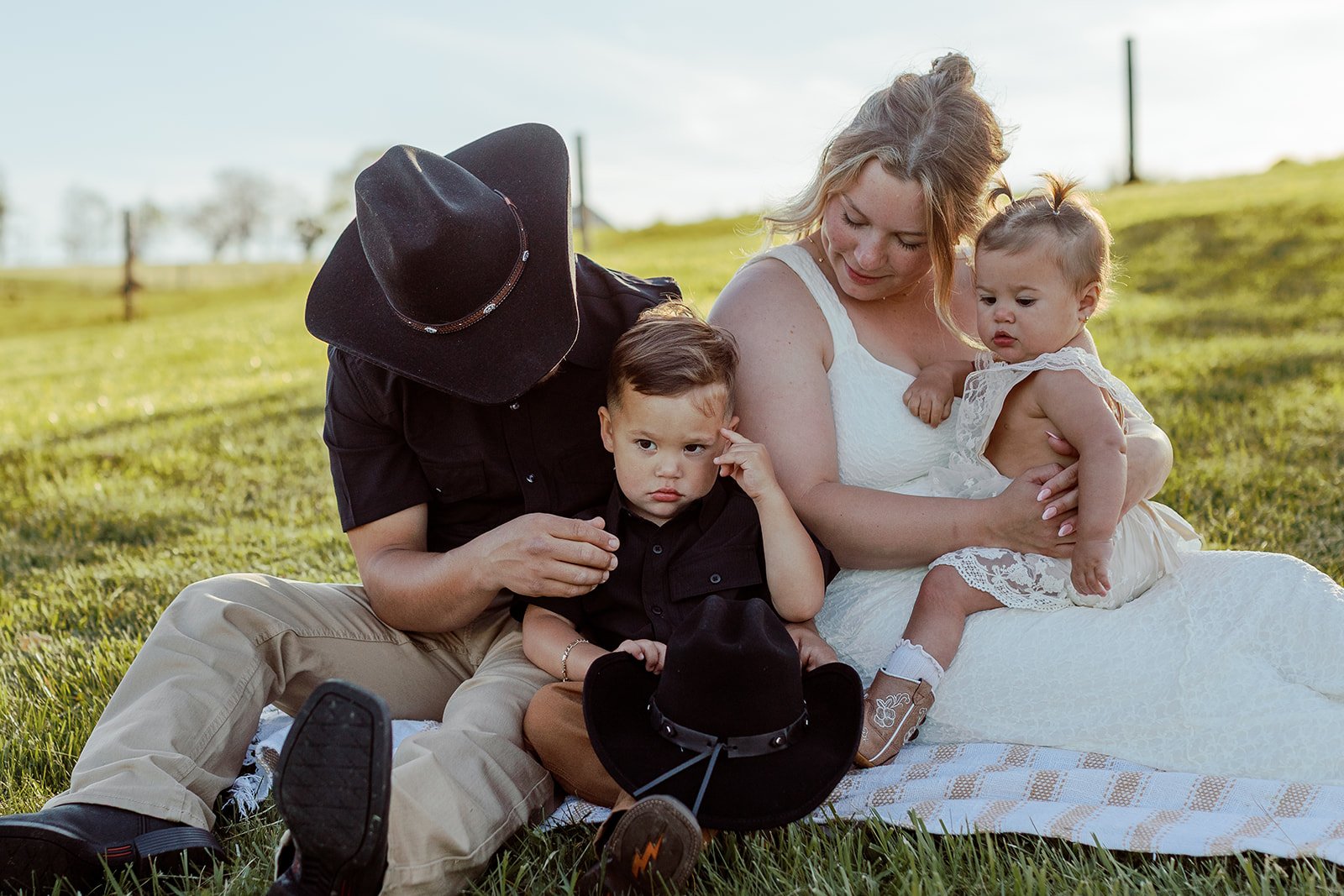 A woman with blonde hair sitting on the grass with three children surrounded by greenery in a park or field on a sunny day, one wearing a white dress, two boys wearing black shirts, and a woman with blonde hair holding a young girl in a white dress.