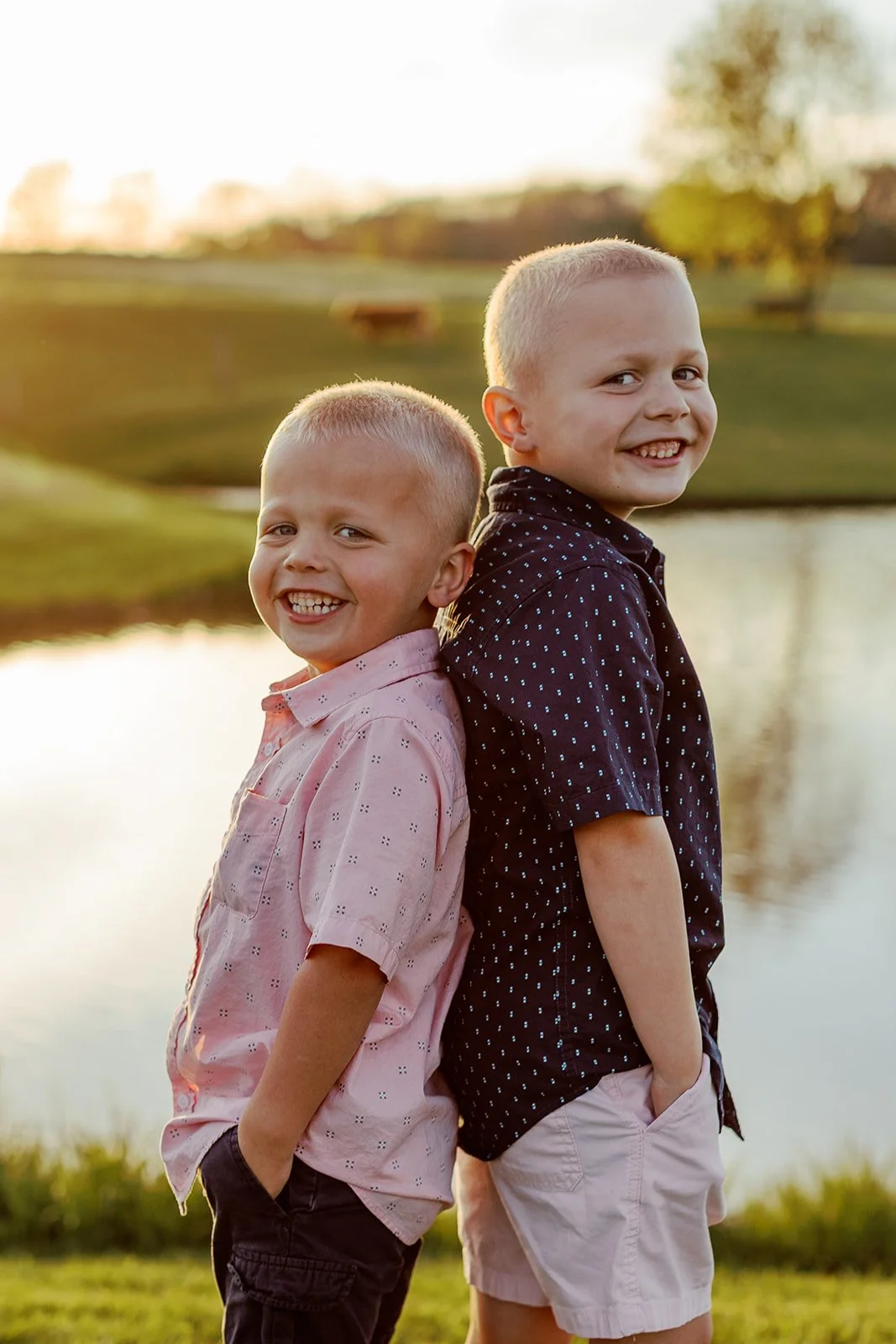 Two young boys standing back-to-back outdoors by a lake during golden hour, smiling at the camera.