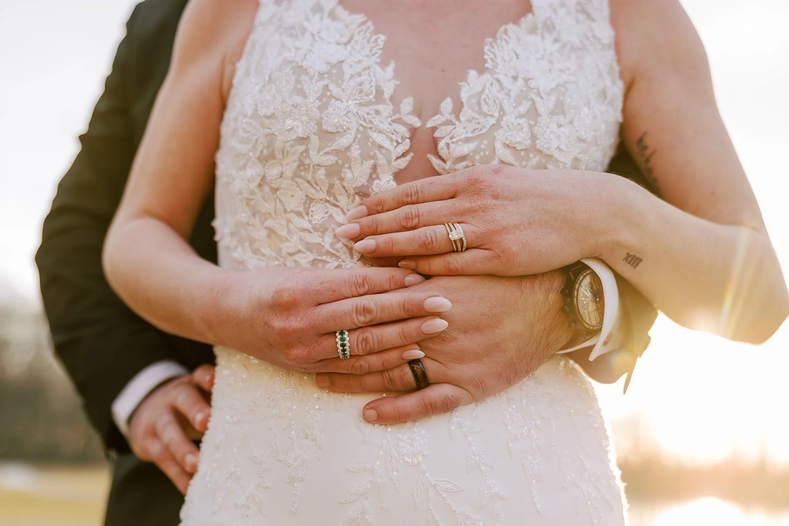Close-up of a bride and groom holding their hands over her stomach during sunset. The bride wears an intricate lace wedding dress and a wedding ring, and the groom wears a black suit, a watch, and a wedding band.