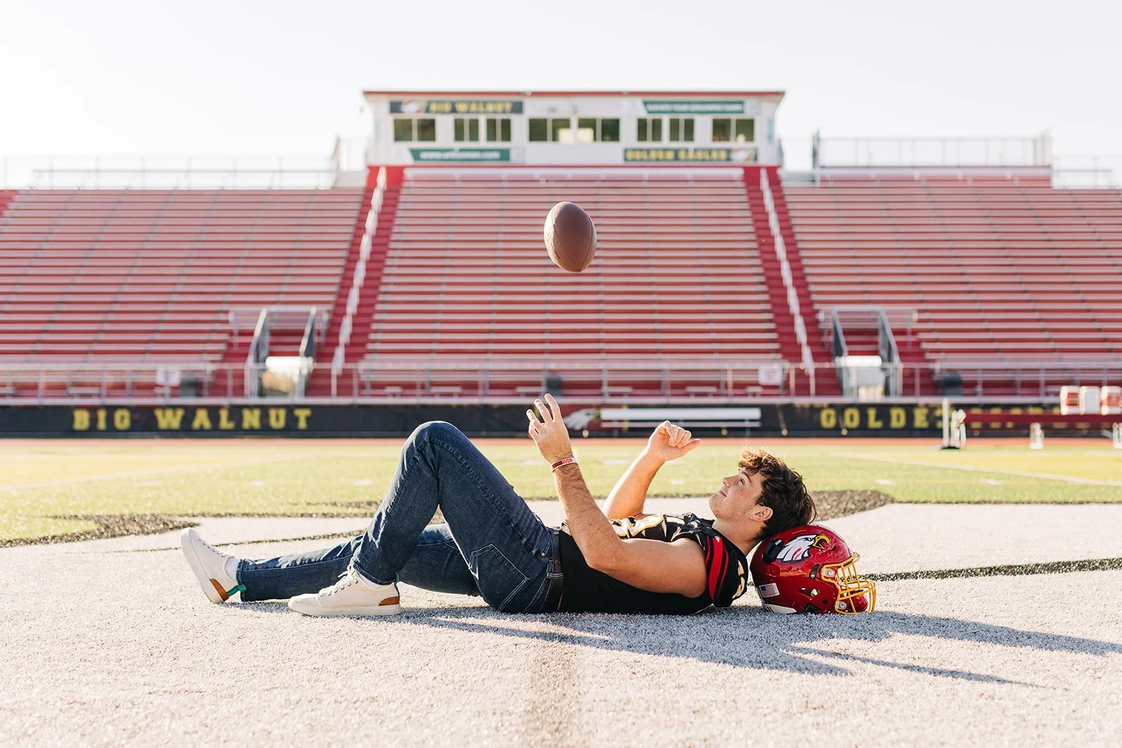 A young man lying on the football field with his helmet beside him, throwing a football in a stadium with empty red bleachers in the background. Senior football session.