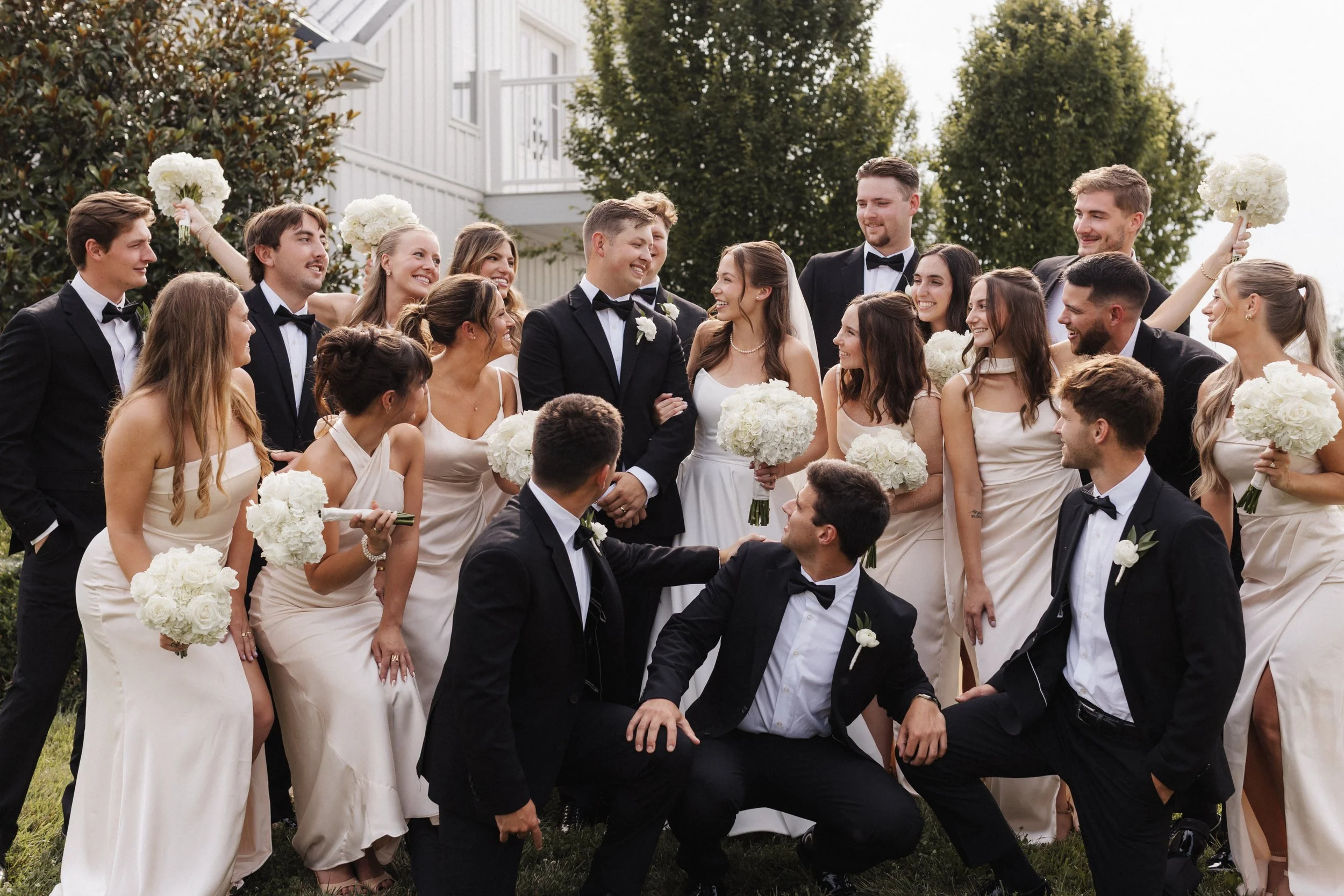 A wedding party outdoors with guests dressed formally. The bride and groom are in the center, smiling at each other. Bridesmaids in beige dresses hold white bouquets, and groomsmen in black tuxedos surround the couple, some holding white flowers. Everyone is smiling and appears joyful.