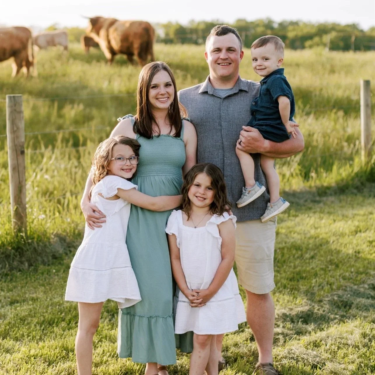 A family of five posing outdoors in a grassy field with bison in the background. The group includes a man, woman, and three young girls, all smiling and dressed in light, summer clothing.