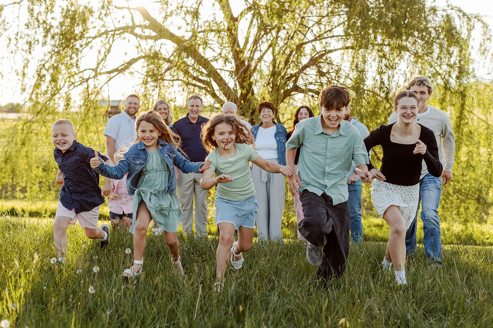 A group of children and adults running and having fun outdoors on a grassy field with a large tree and green landscape in the background, during sunset. Extended family session.