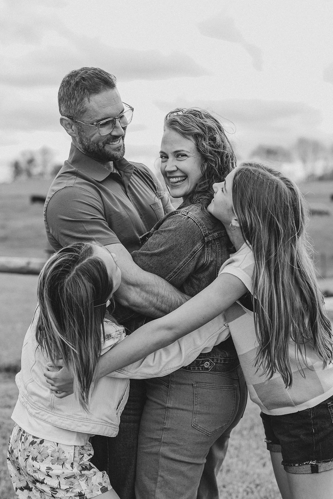 A family of four hugging outdoors, black and white photo, smiling and enjoying each other's company.