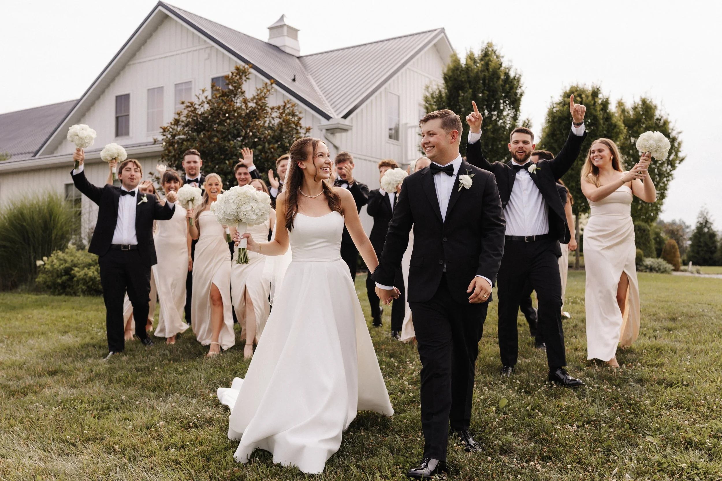 A bride and groom walking together during a wedding celebration with guests in the background, holding bouquets, outside a white house with a gabled roof and trees. Wedding photographer. Magnolia Hill Farm.