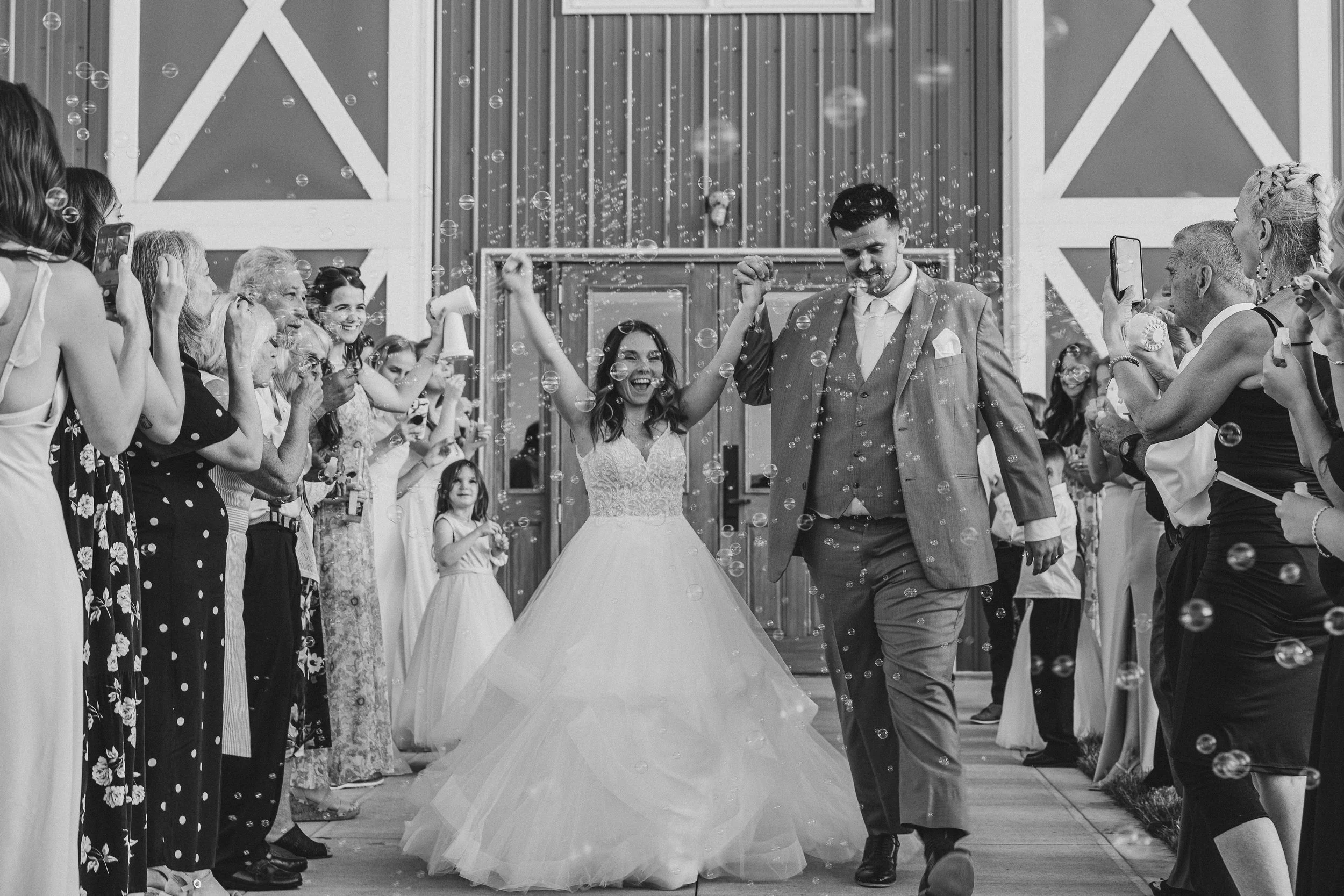 A bride and groom walking hand in hand through a crowd of guests at their wedding reception, surrounded by bubbles and smiling faces.