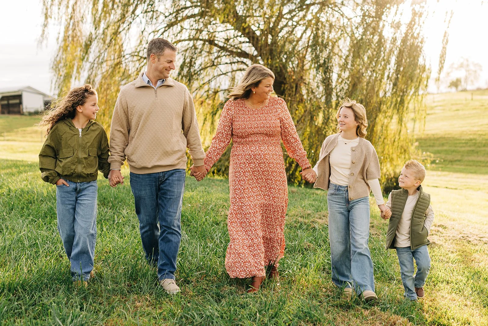 A family of six walking outdoors holding hands, smiling and enjoying time together on a sunny day with a large tree in the background. Family photographer.