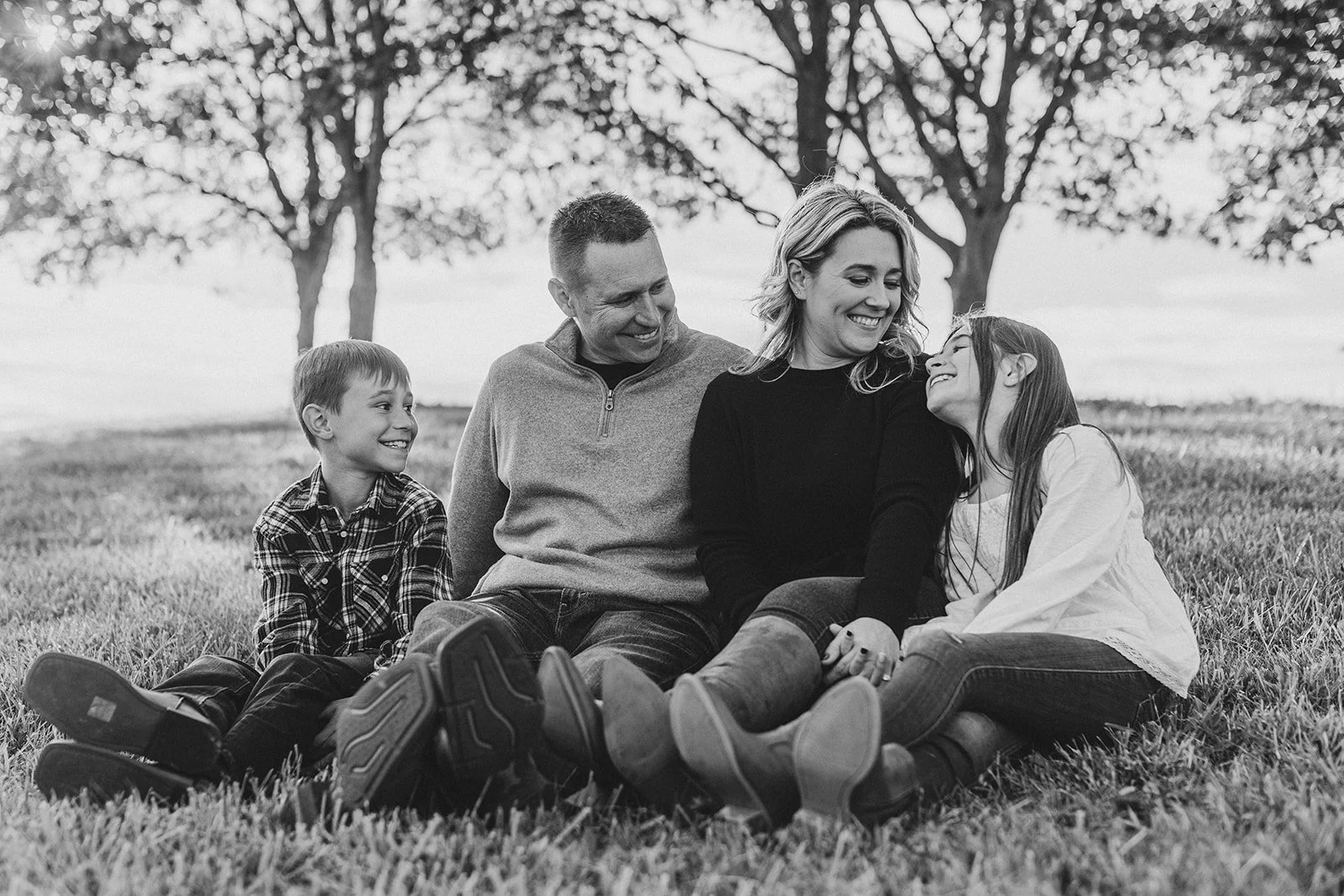 A black and white photograph of a family sitting on the grass in a park with trees in the background, smiling and enjoying each other's company.